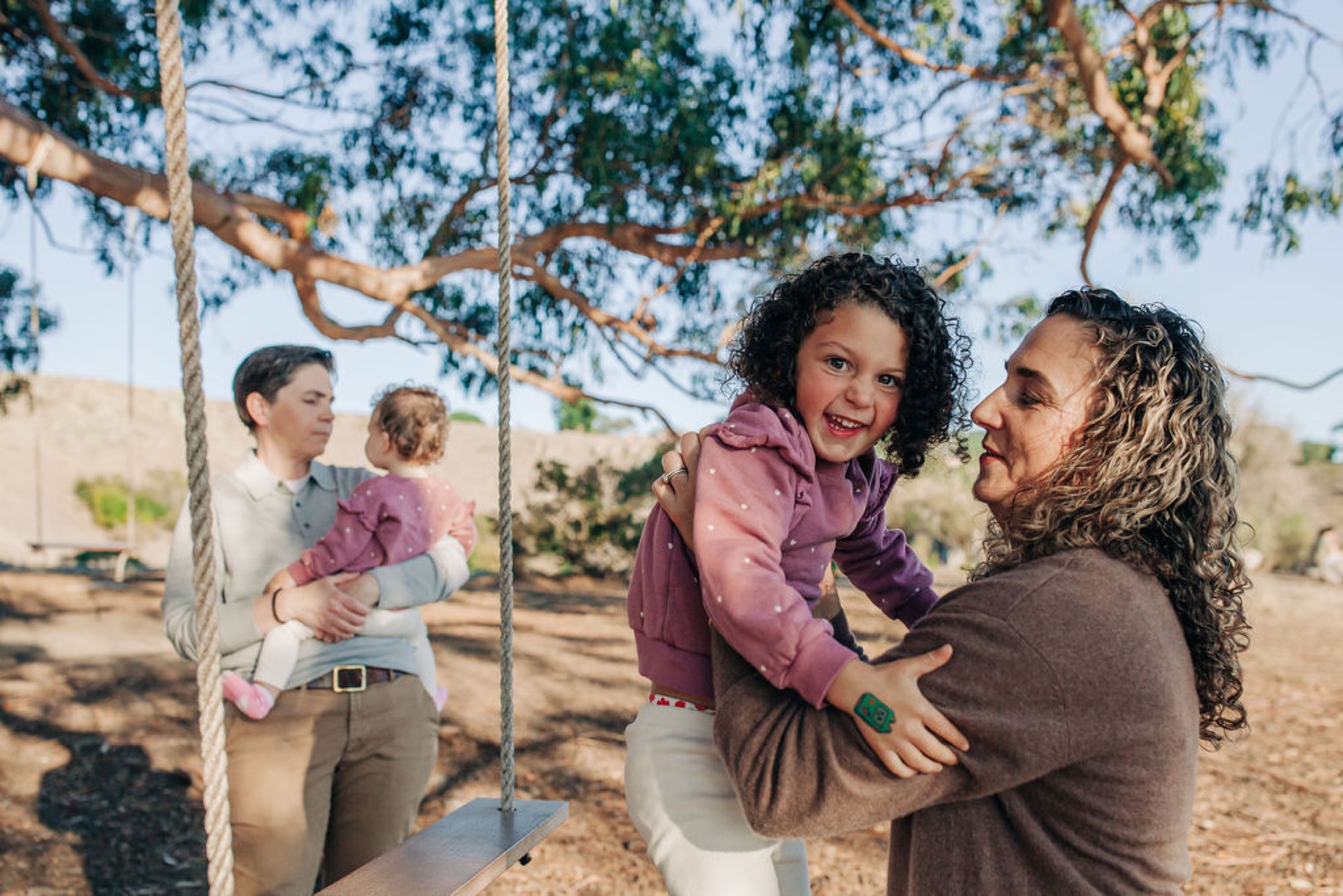 A man with curly hair lifts a laughing girl on a rope swing while another child watches nearby under a sprawling eucalyptus tree