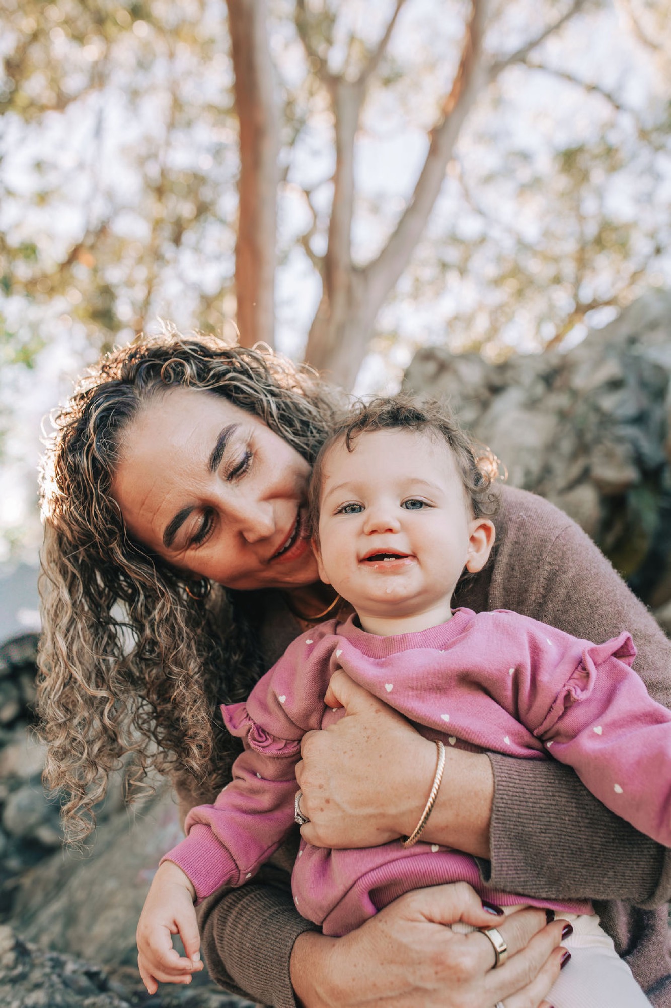 Mother with closed eyes gently kisses toddler's cheek beneath blooming tree, soft golden light filtering through canopy