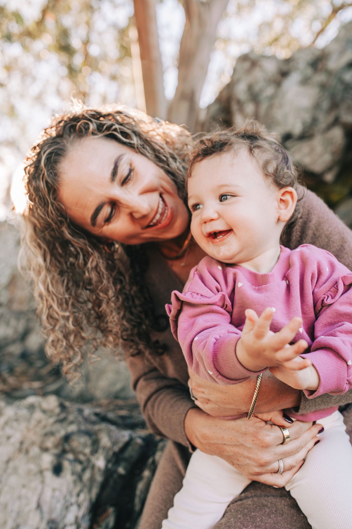 A mother with curly hair and warm smile holds her laughing toddler in a pink hoodie beneath dappled sunlight filtering through tree branches