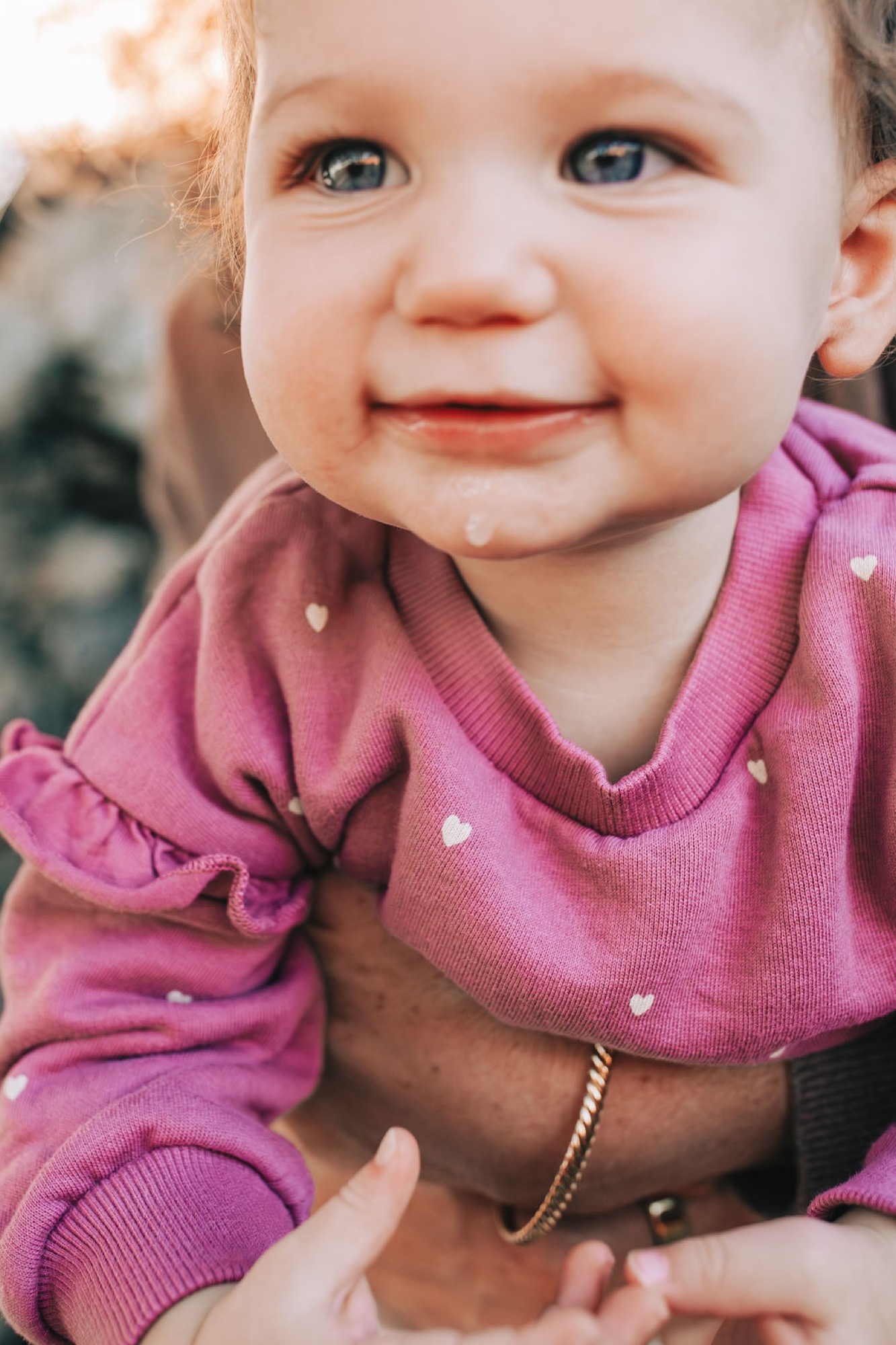 A toddler in a pink polka-dot sweater beams with a genuine smile while held by an adult wearing a gold bracelet, backlit by soft golden hour light