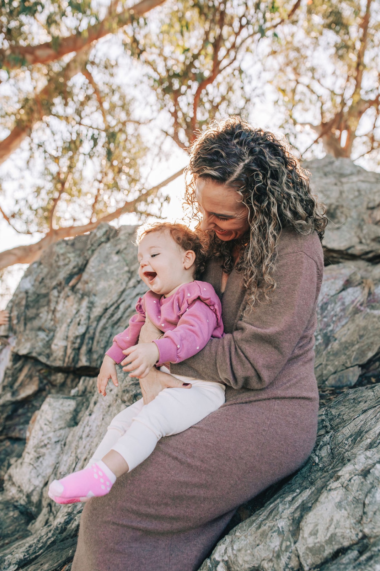 A mother with curly hair holds her laughing toddler on a weathered rock beneath a sprawling tree with dappled sunlight filtering through