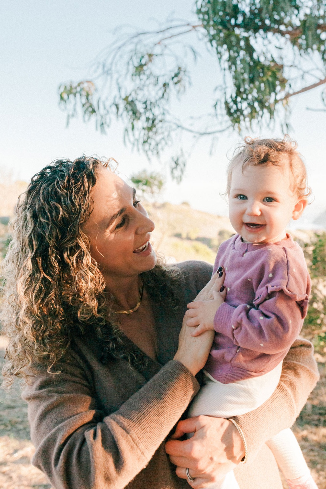 Mother and toddler laugh together under snow-laden eucalyptus branches, golden afternoon light warming their faces on the hillside