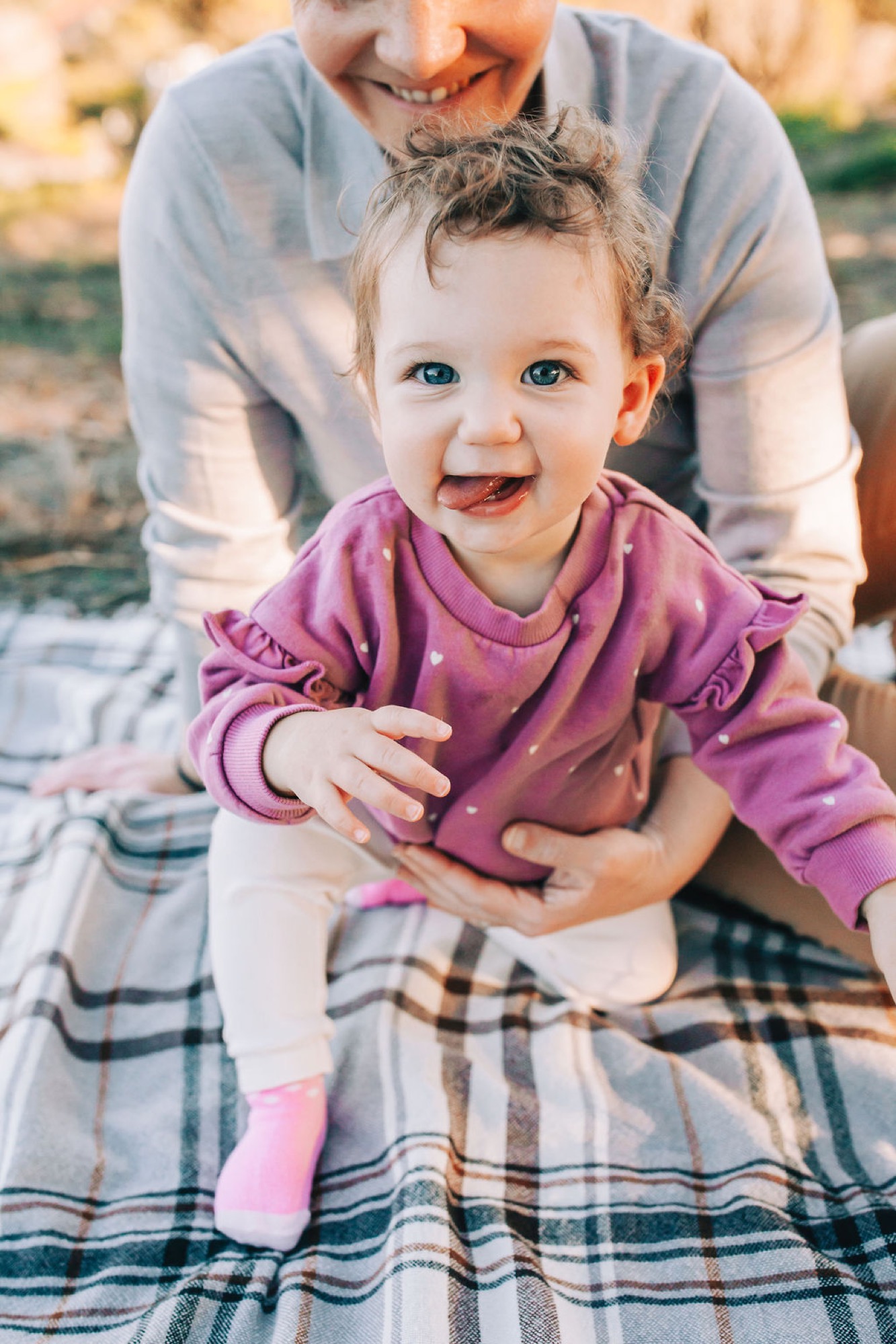 Beaming toddler in magenta sweatshirt sits on plaid blanket while parent holds her, golden afternoon light illuminating her bright blue eyes and joyful expression