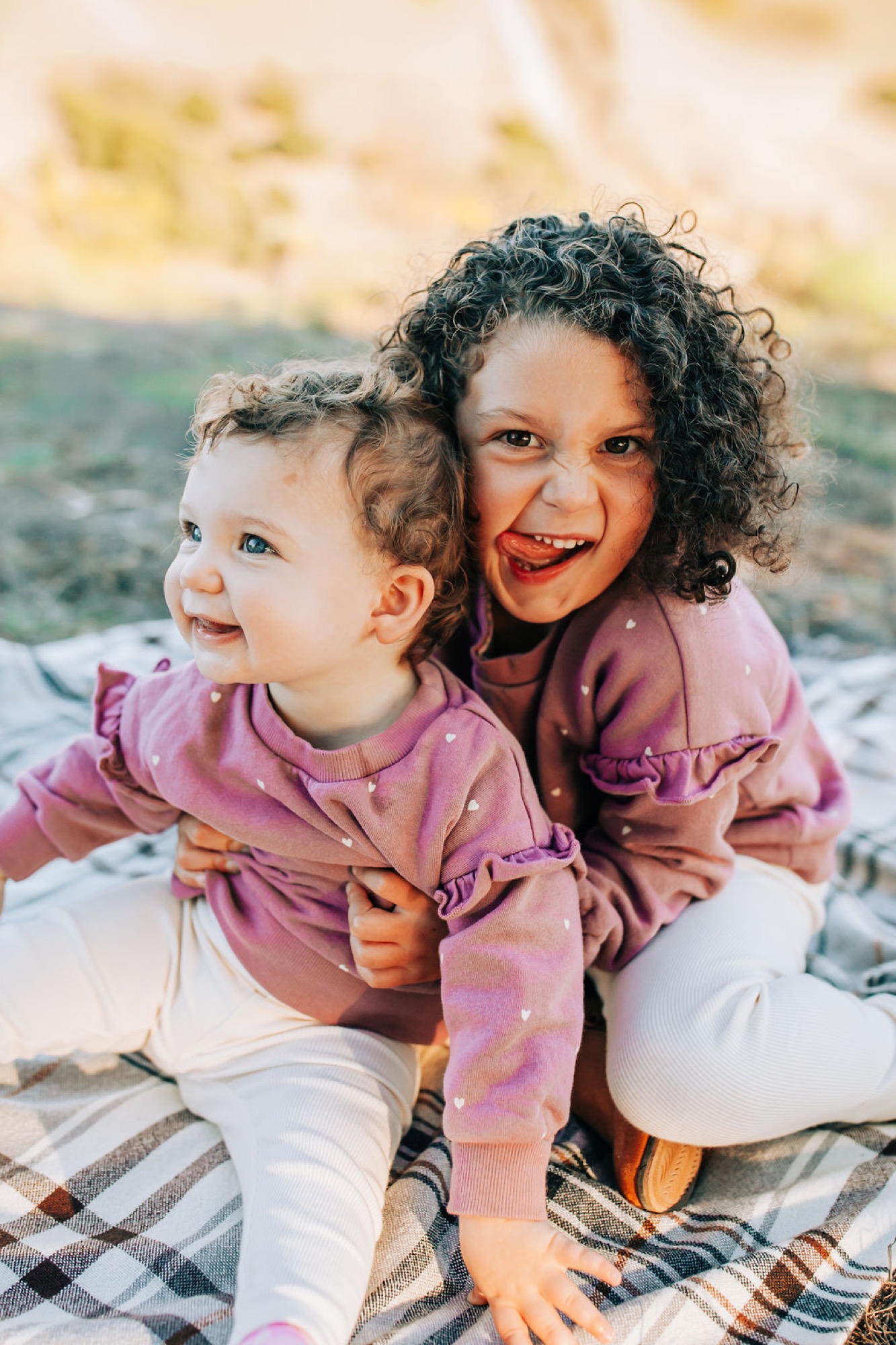 Two sisters with curly dark hair beam with genuine joy while sitting together on a plaid blanket, their matching pink polka-dot tops creating visual harmony against the sandy beach backdrop