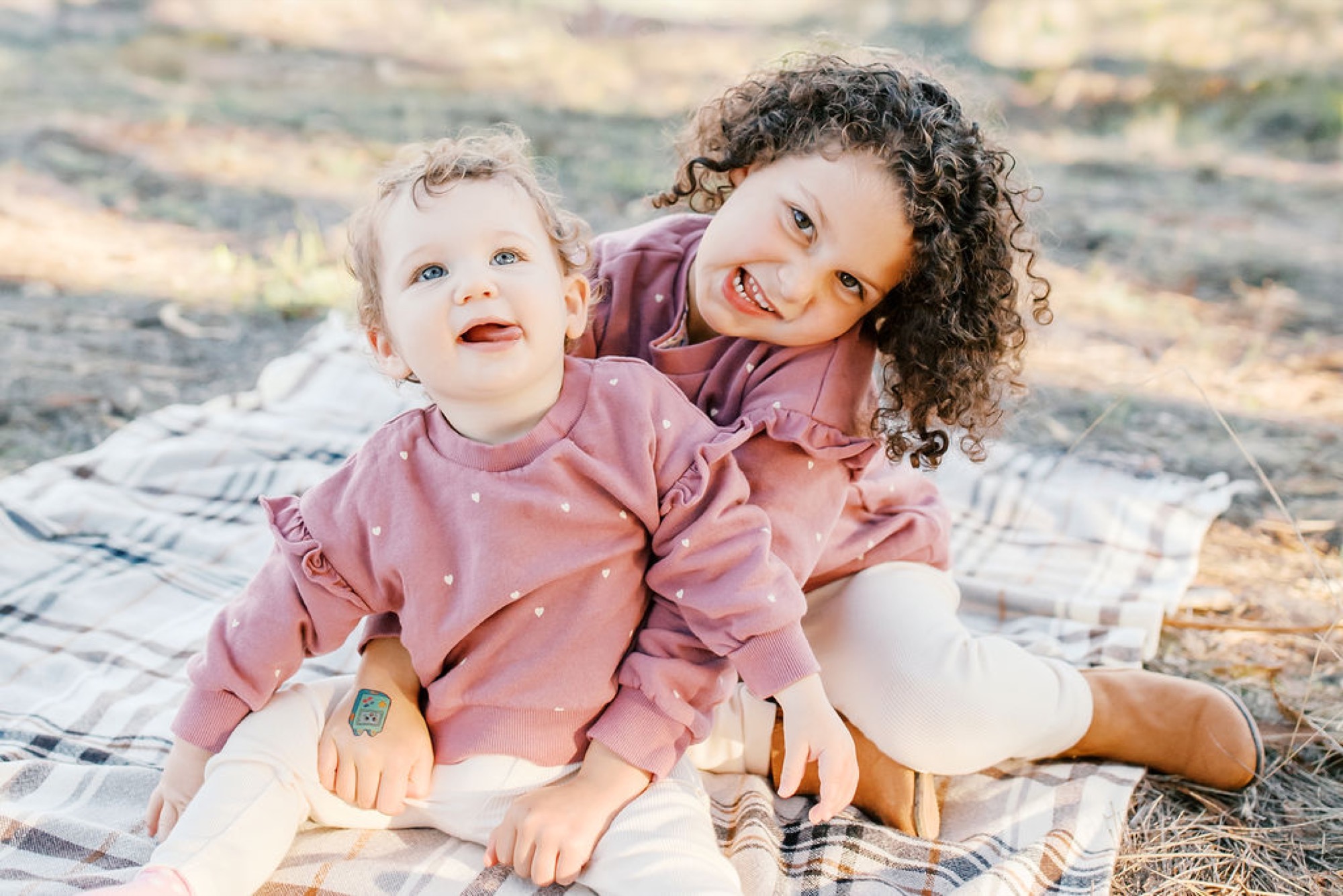 Two young girls in matching pink polka-dot sweatshirts sit on a plaid blanket, the older child beaming while cradling her toddler sibling on rocky terrain