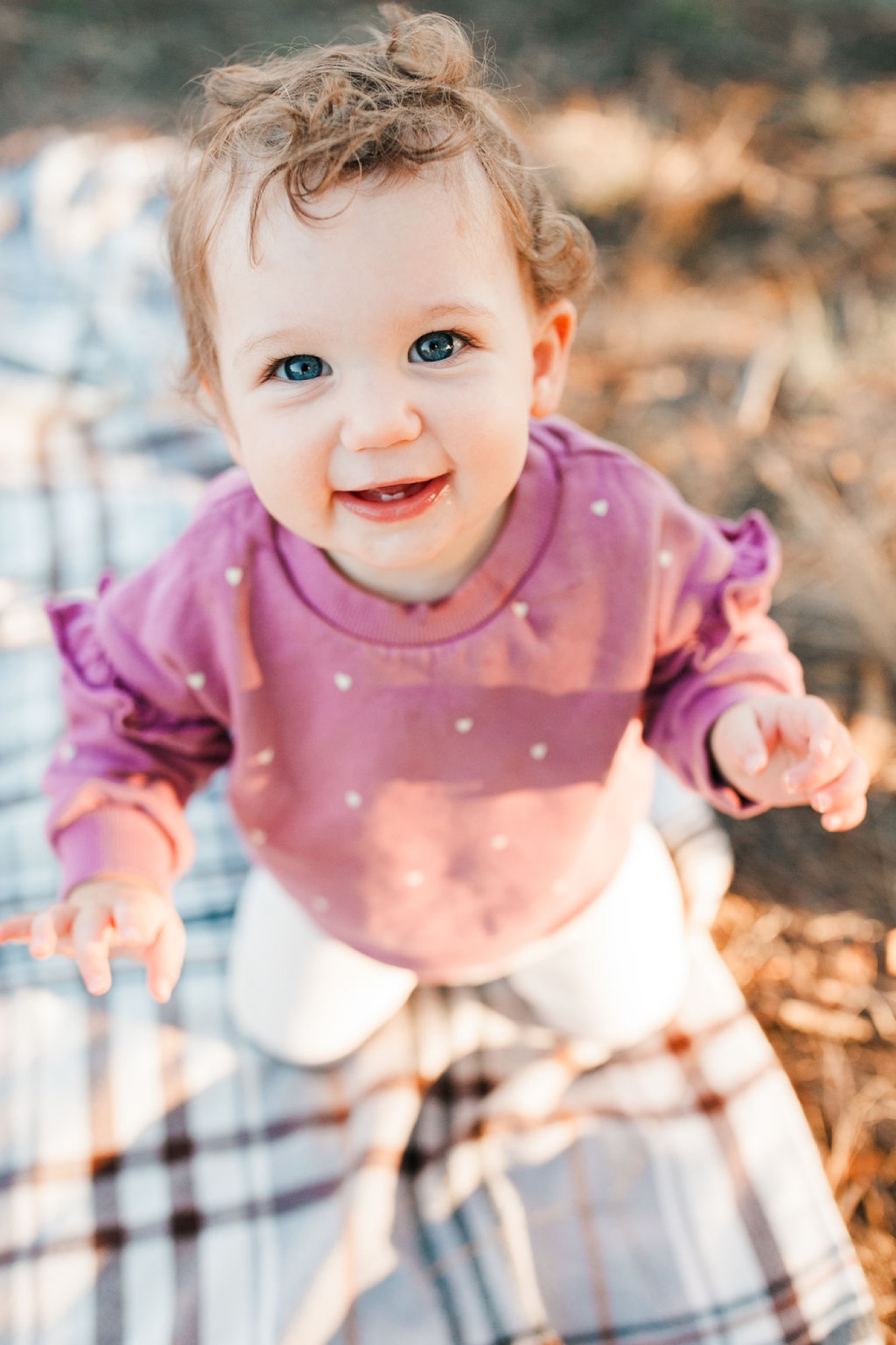 Toddler in purple polka-dot shirt beams with open-mouthed joy while crawling toward camera on plaid blanket, golden-hour sunlight illuminating her bright blue eyes