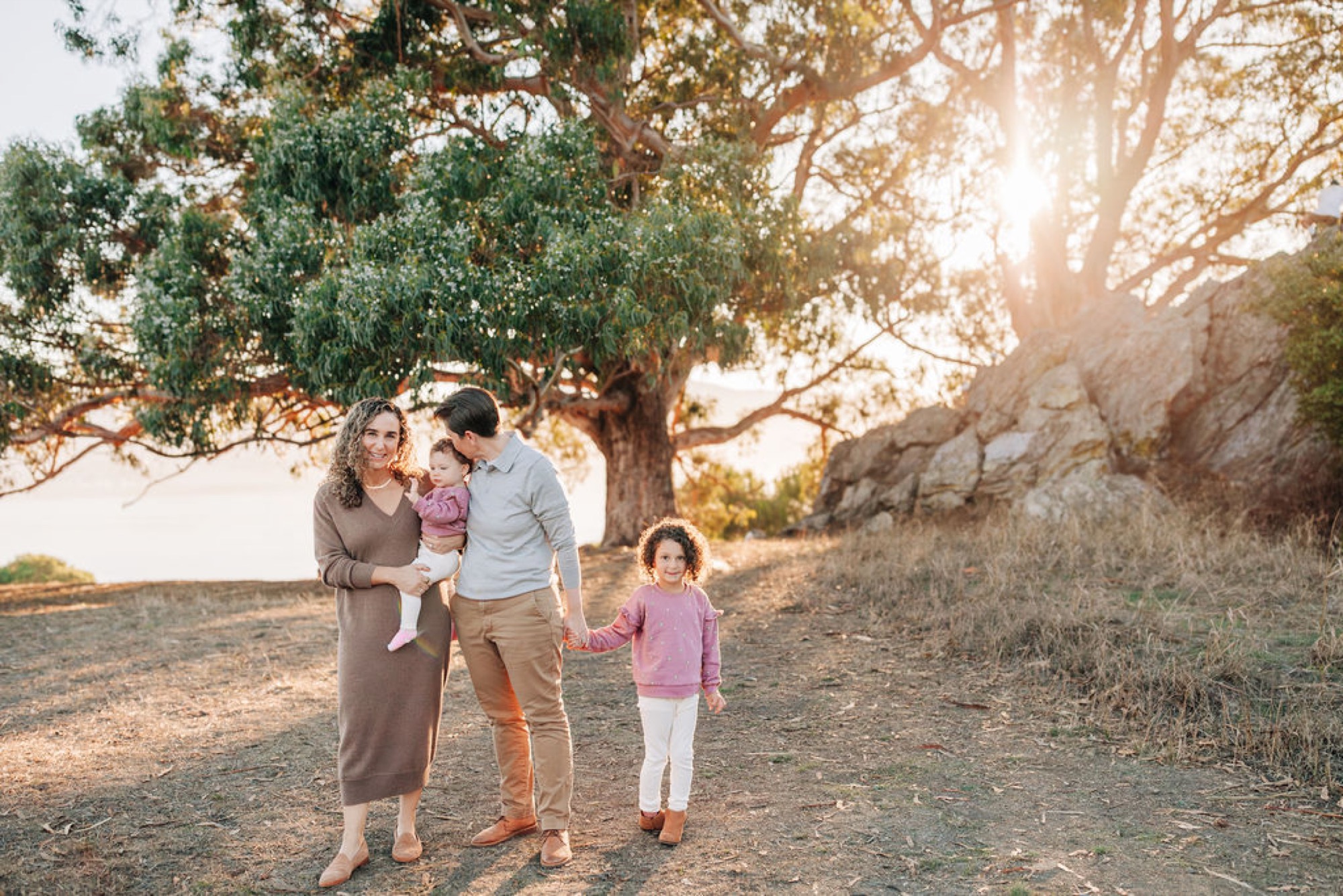 Family of four walks hand-in-hand beneath a sprawling oak tree during golden hour, soft light filtering through the branches