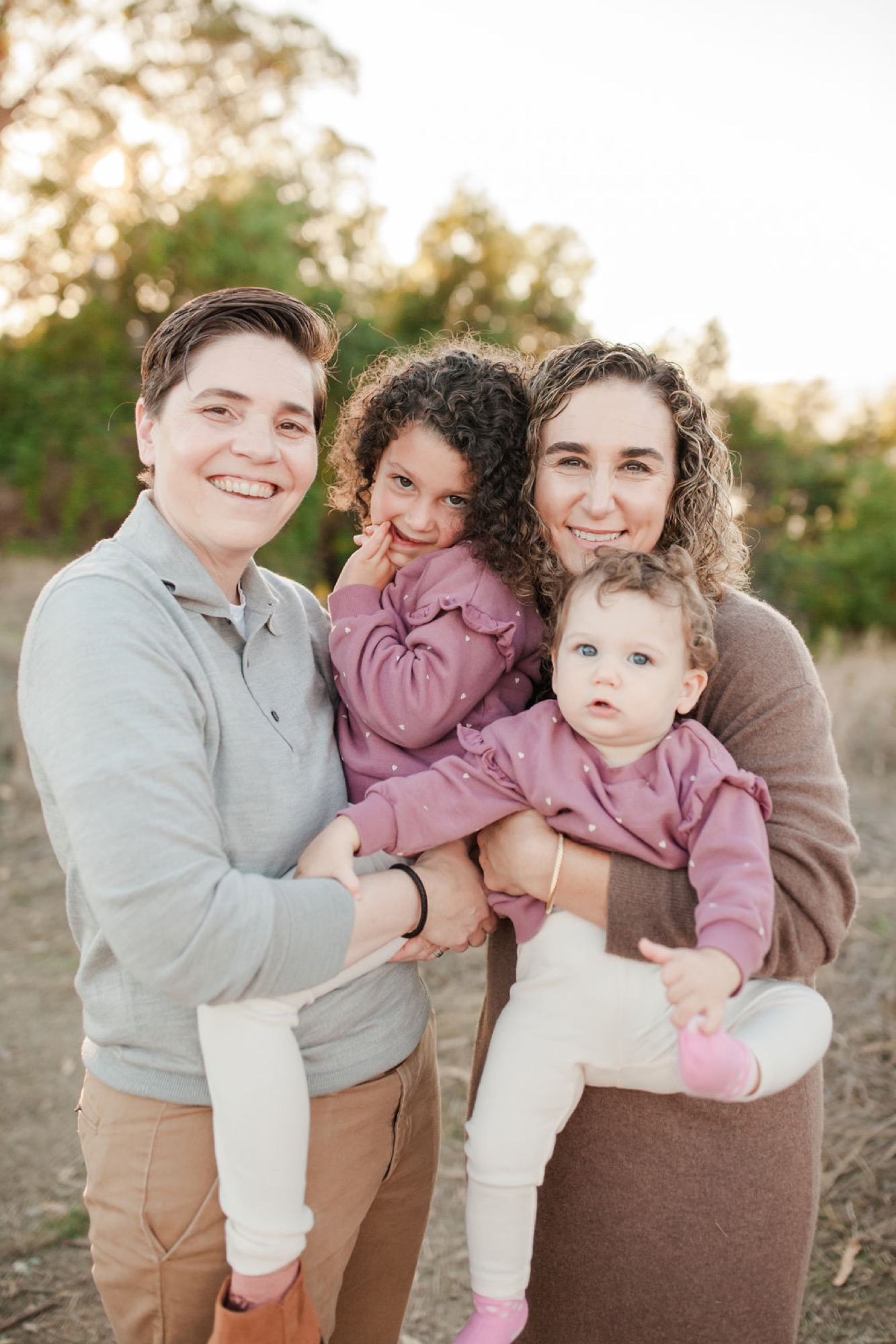 Two adults and two young children smile warmly at the camera on a sunlit path, with blooming white flowers softly glowing in the background