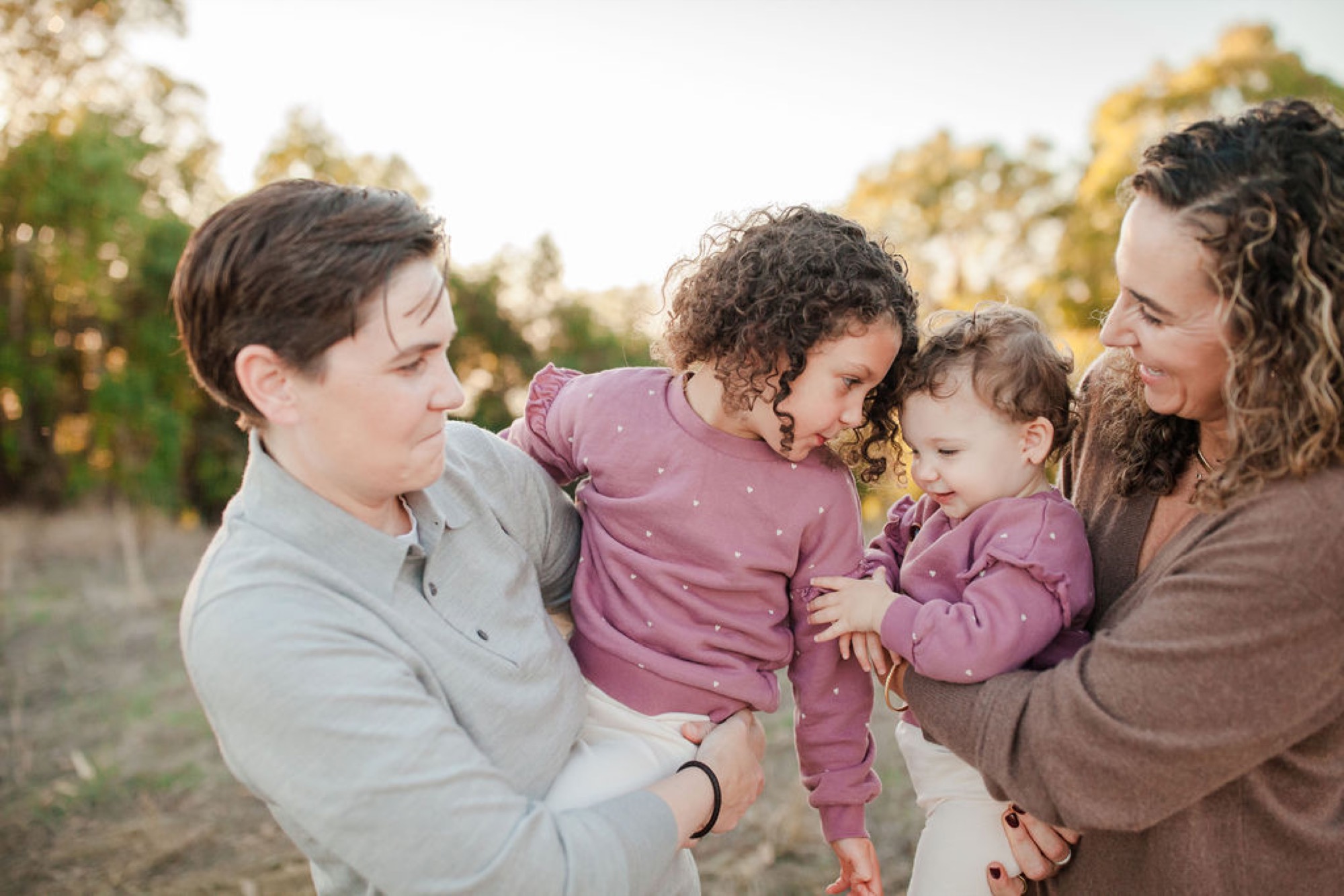 Two adults and two young children in matching mauve outfits share a joyful moment together, golden afternoon light filtering through the forest behind them