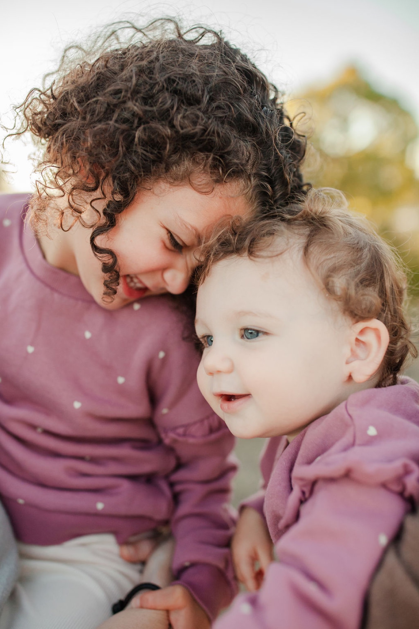 Two curly-haired toddlers in matching mauve polka-dot outfits share a tender moment, with the older child gazing down affectionately at the younger one outdoors