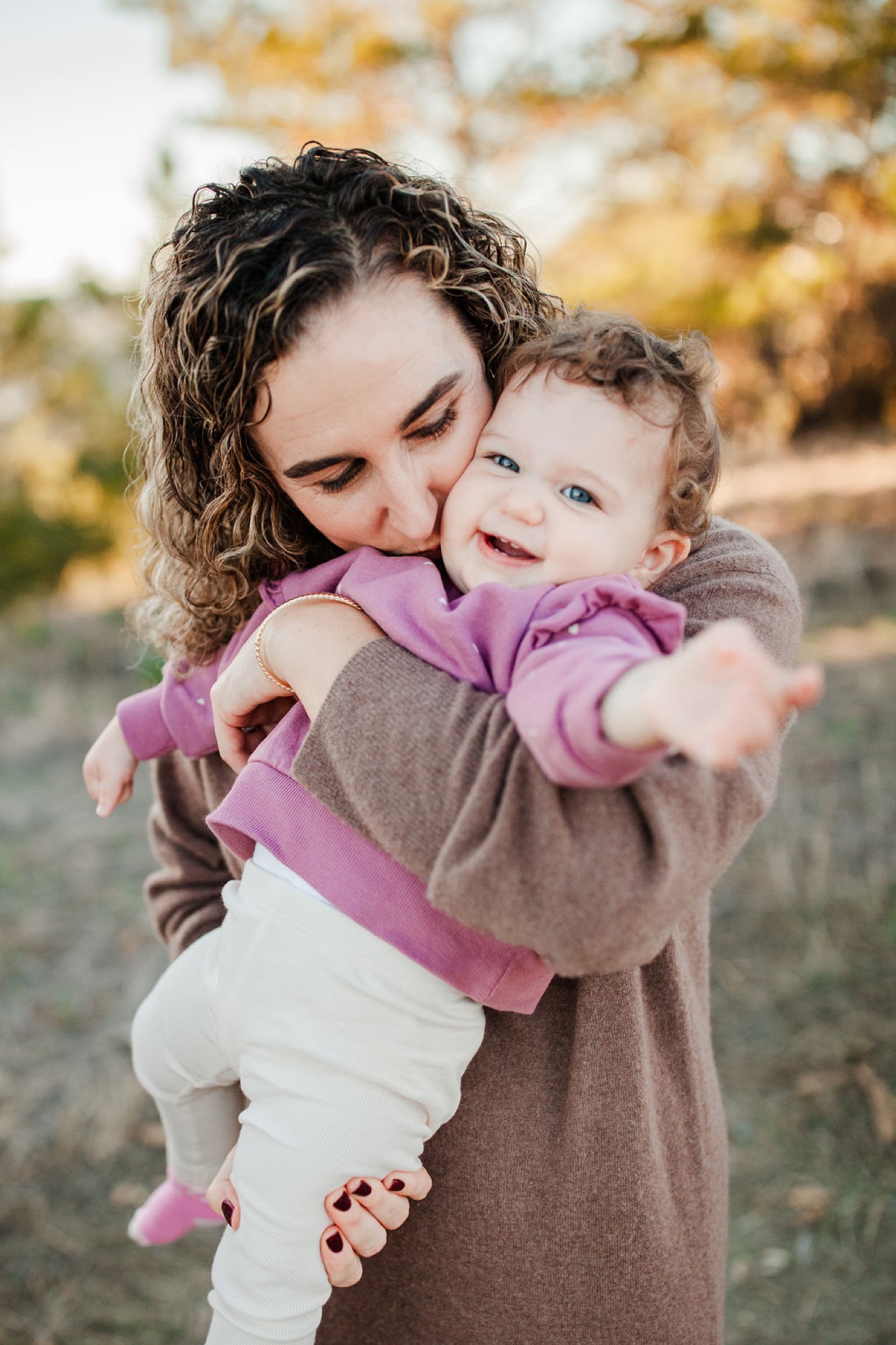 Mother holds her toddler aloft with genuine affection, both beaming with joy against warm autumn foliage and soft natural light