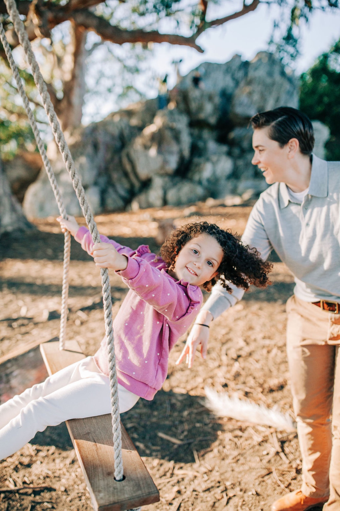 A curly-haired toddler beams while gripping a rope swing as her mother watches with genuine warmth beneath dappled tree shade
