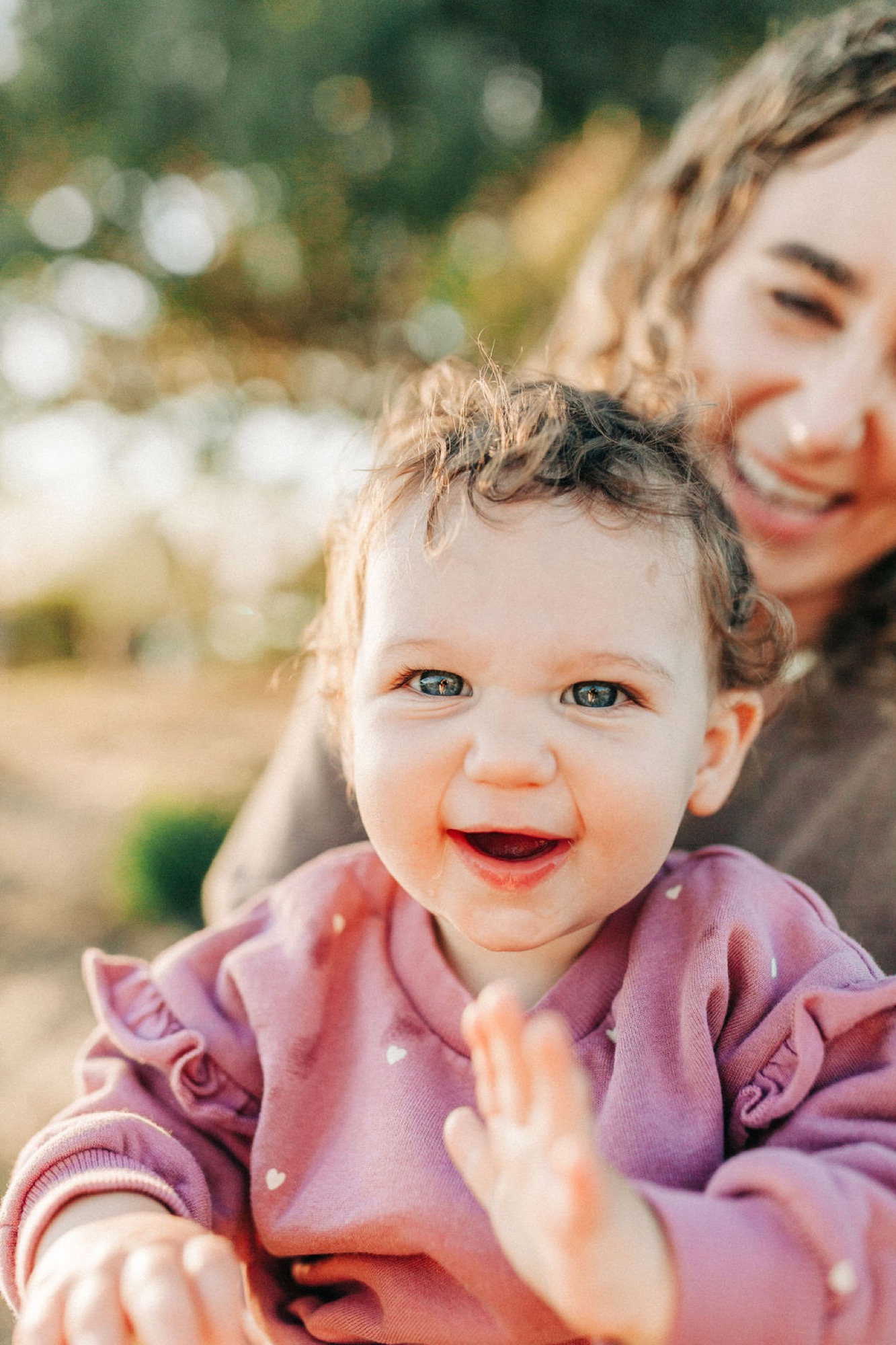 Toddler in mauve ruffled dress beams with pure joy while held by parent, golden afternoon light catching her bright eyes and curly hair