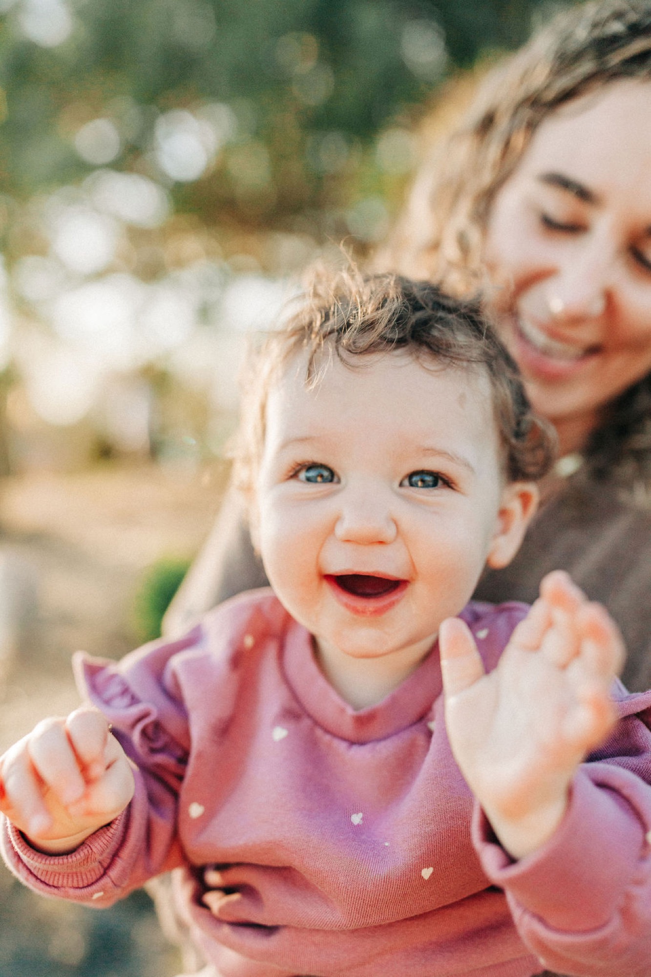 Toddler in pink laughs with open mouth while mother holds her close in dappled sunlight, both radiating joy