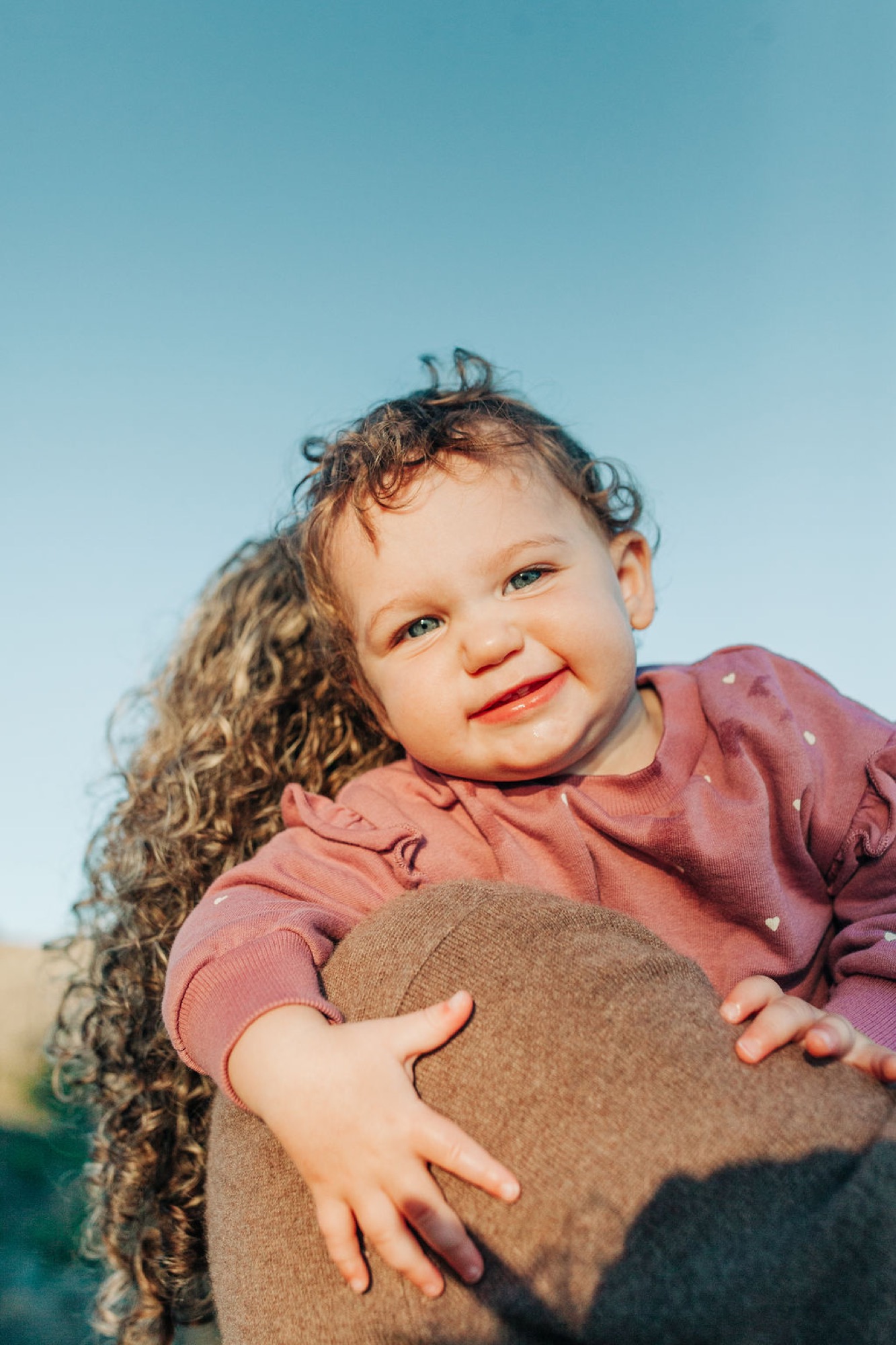 Toddler with curly hair and bright smile gazes at camera while held against parent's chest in golden afternoon light