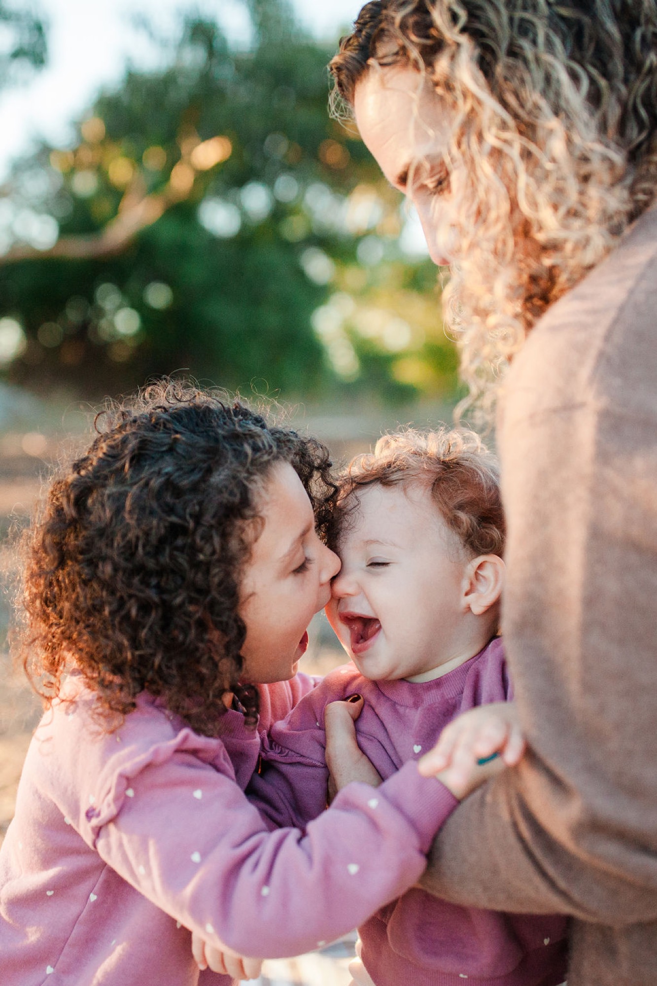 A mother kisses her laughing toddler while a family member watches, golden afternoon light illuminating their joyful outdoor moment