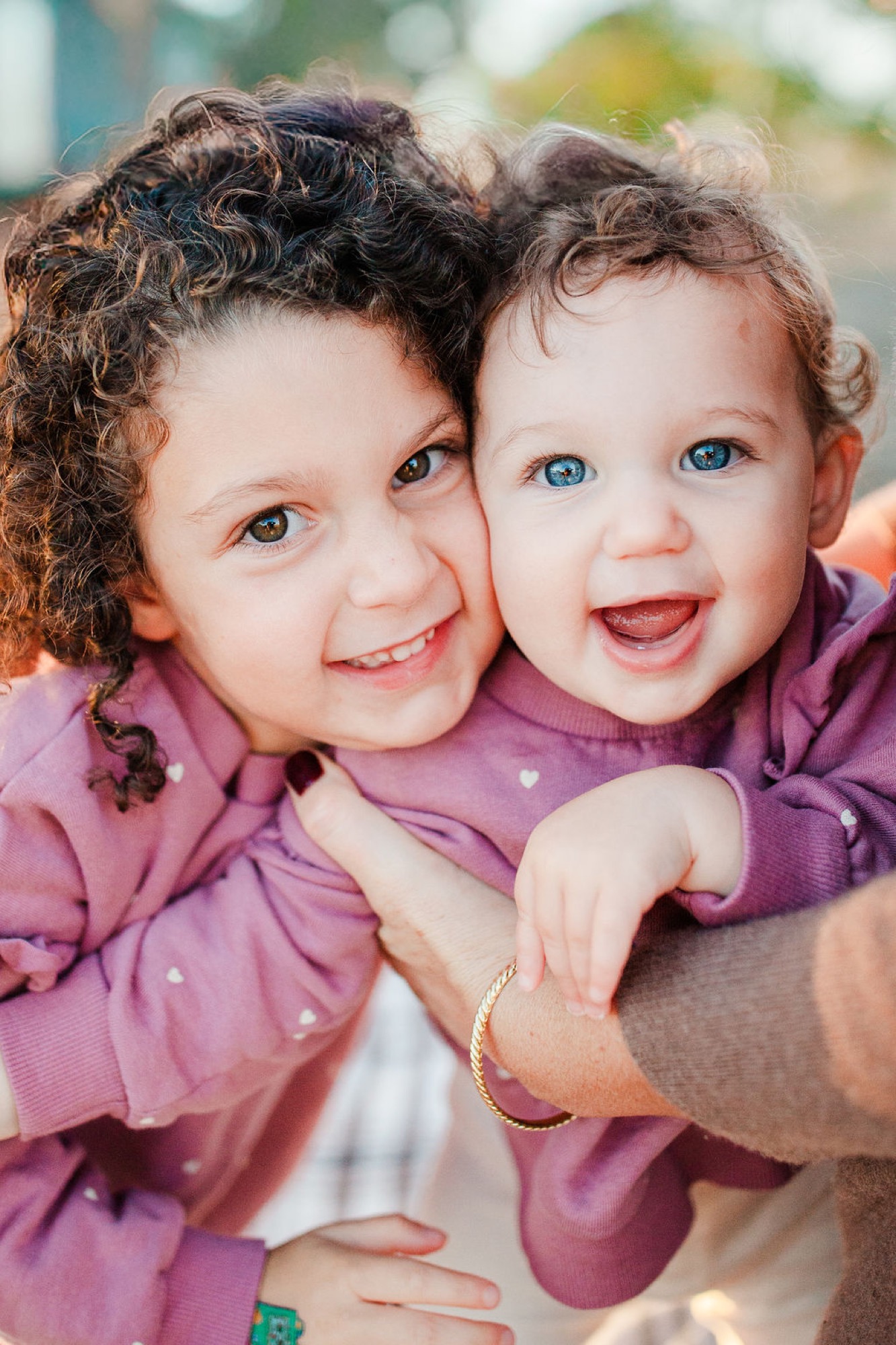 Two young children in matching purple shirts beam with infectious joy, their bright expressions and close embrace radiating warmth in soft outdoor light