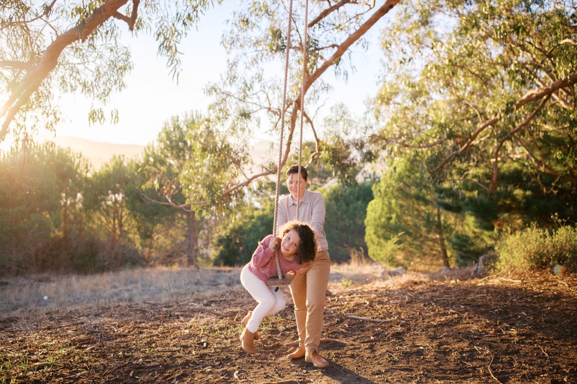 A young girl and man swing together on a rope suspended from a sprawling eucalyptus tree, golden afternoon light filtering through the canopy behind them