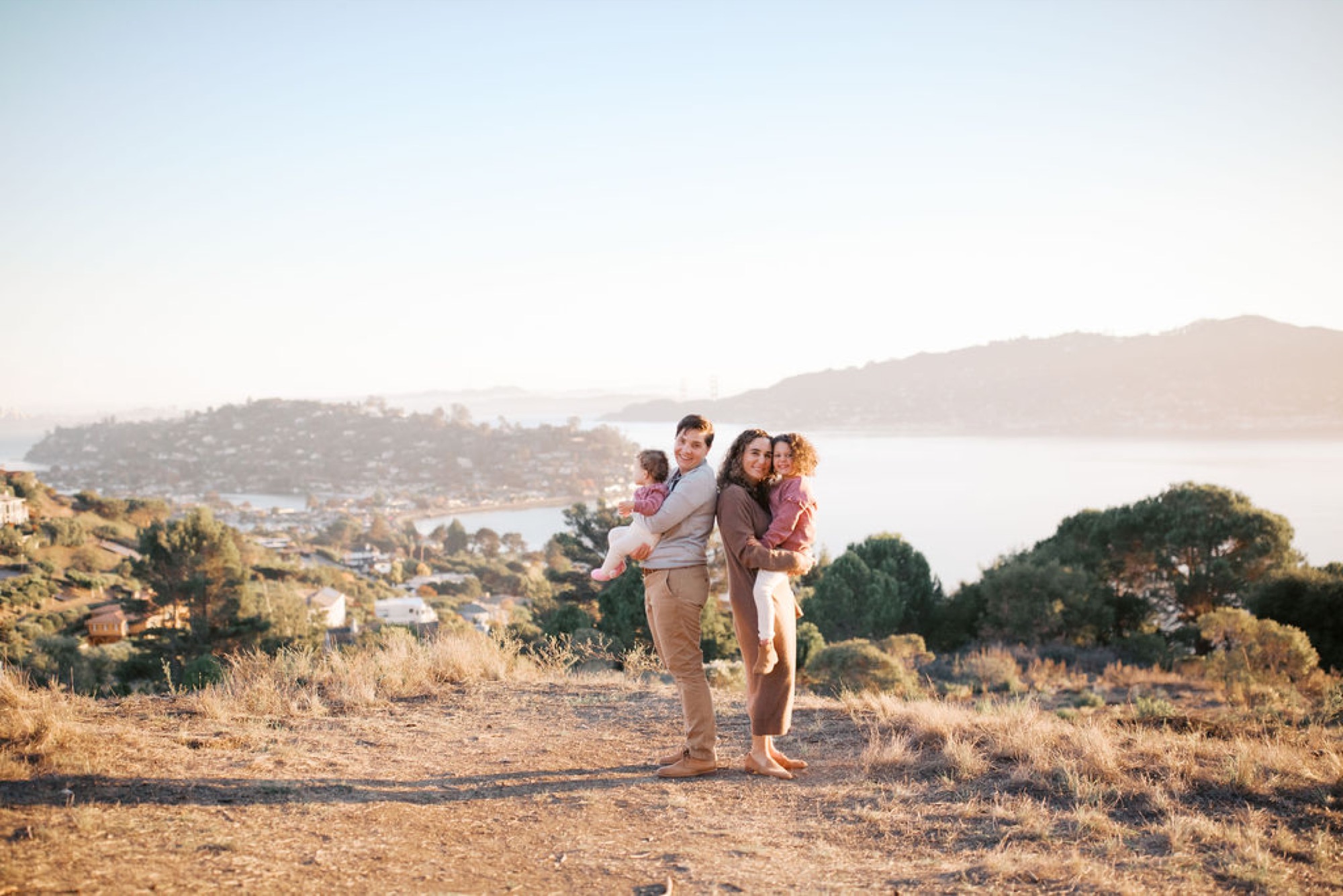 Parents hold their two young daughters on a dusty hilltop trail, backlit by golden afternoon sun with the misty bay and hillside homes visible below