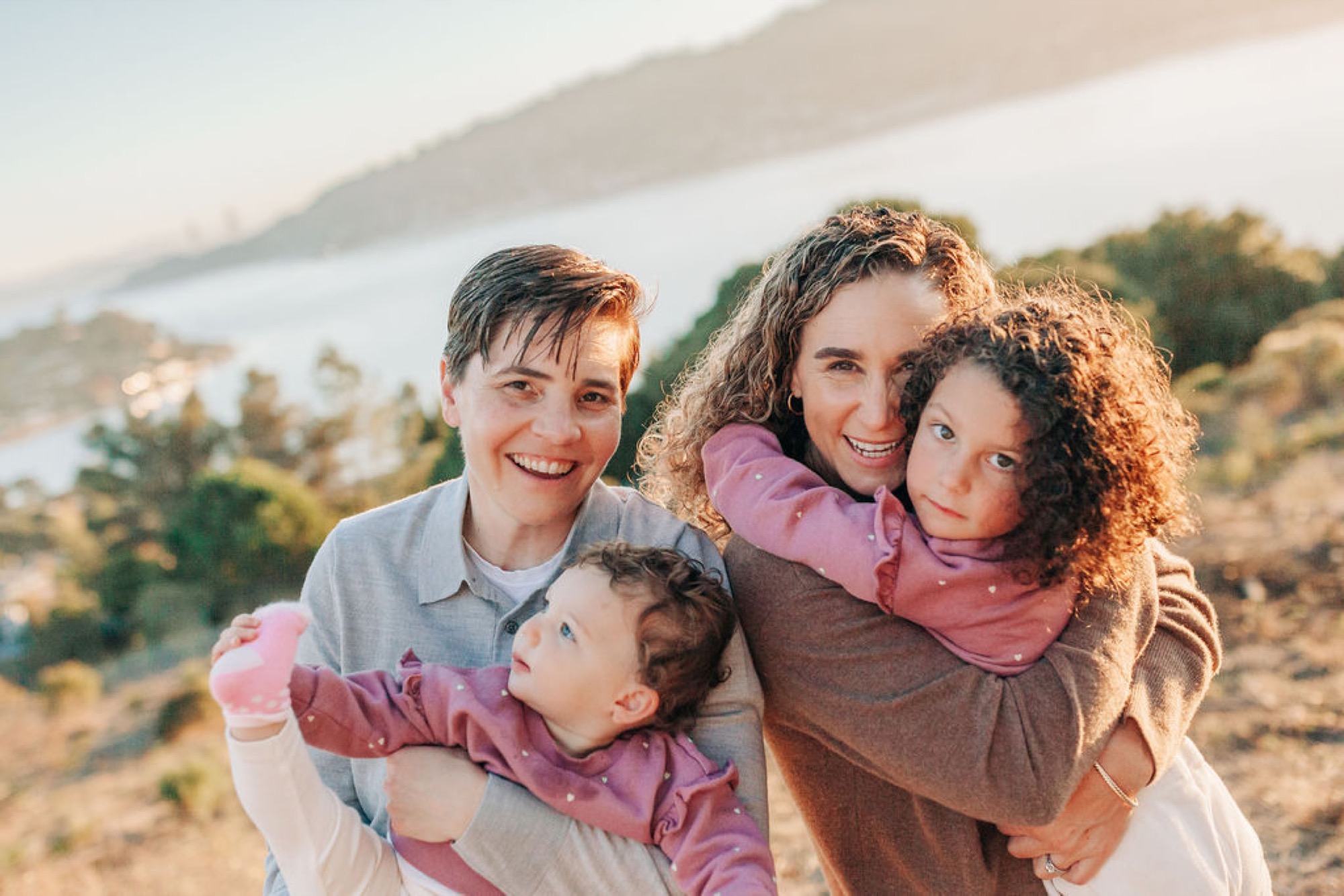 A joyful family of four beams at golden hour on a hillside overlooking the bay, warm sunlight illuminating their genuine smiles
