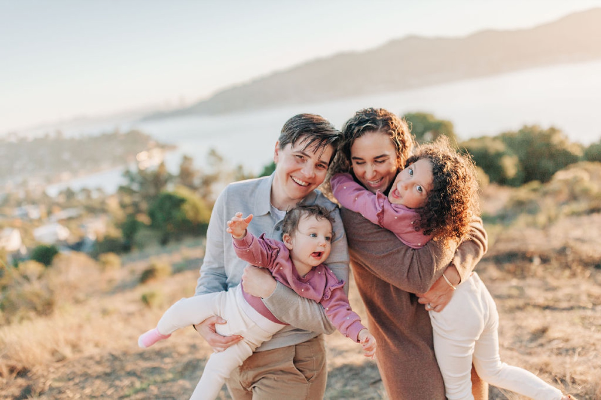 Two parents laugh joyfully while holding their two young children on a sunlit hillside overlooking a distant bay and forested landscape