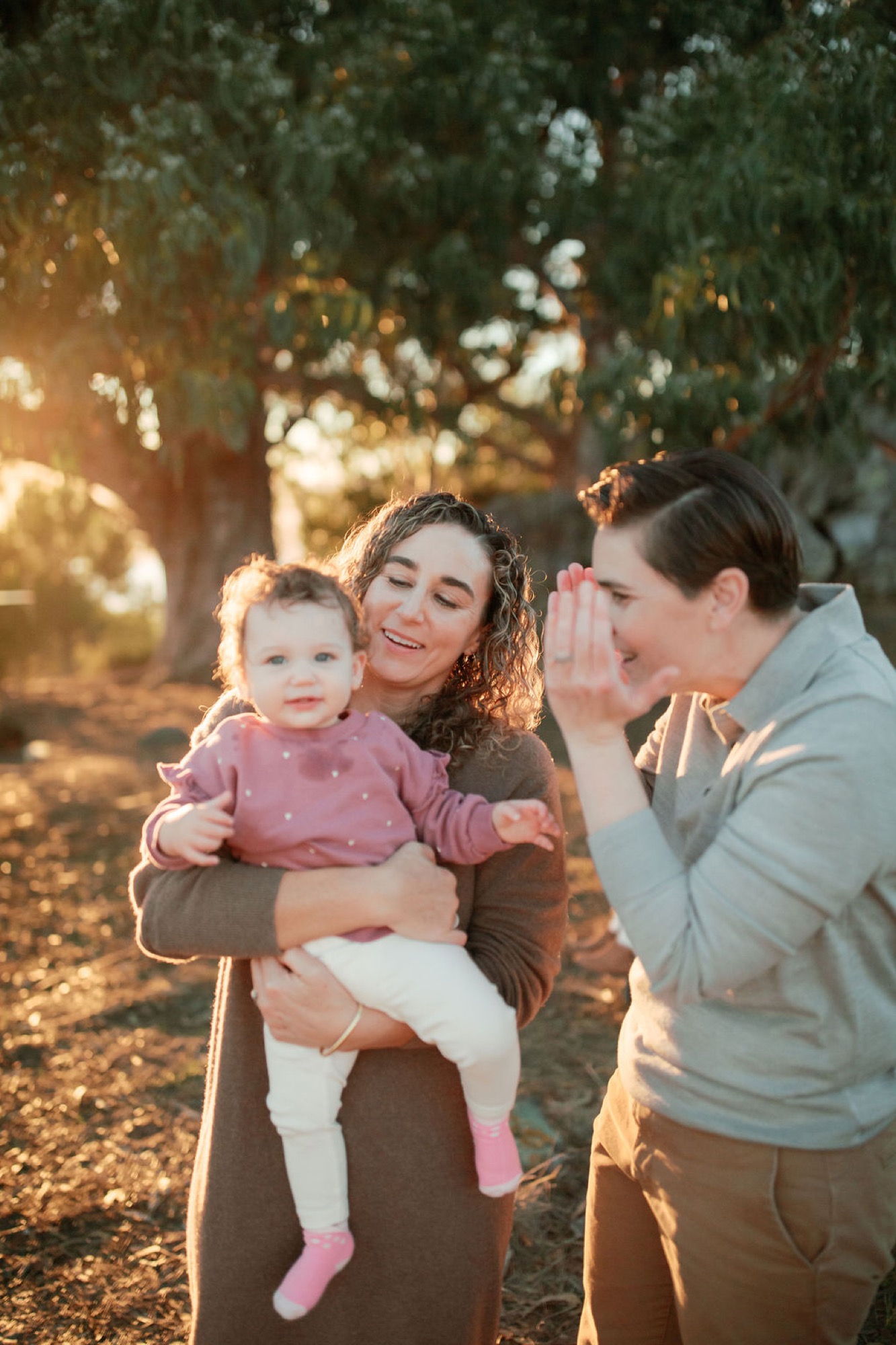 Two adults laugh together while one holds a toddler in pink, bathed in golden sunset light beneath eucalyptus trees