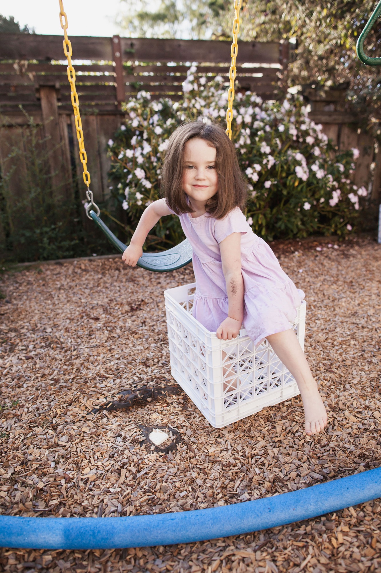 Girl in lavender dress sits confidently on white crate swing, soft afternoon light filtering through blooming flowers behind her