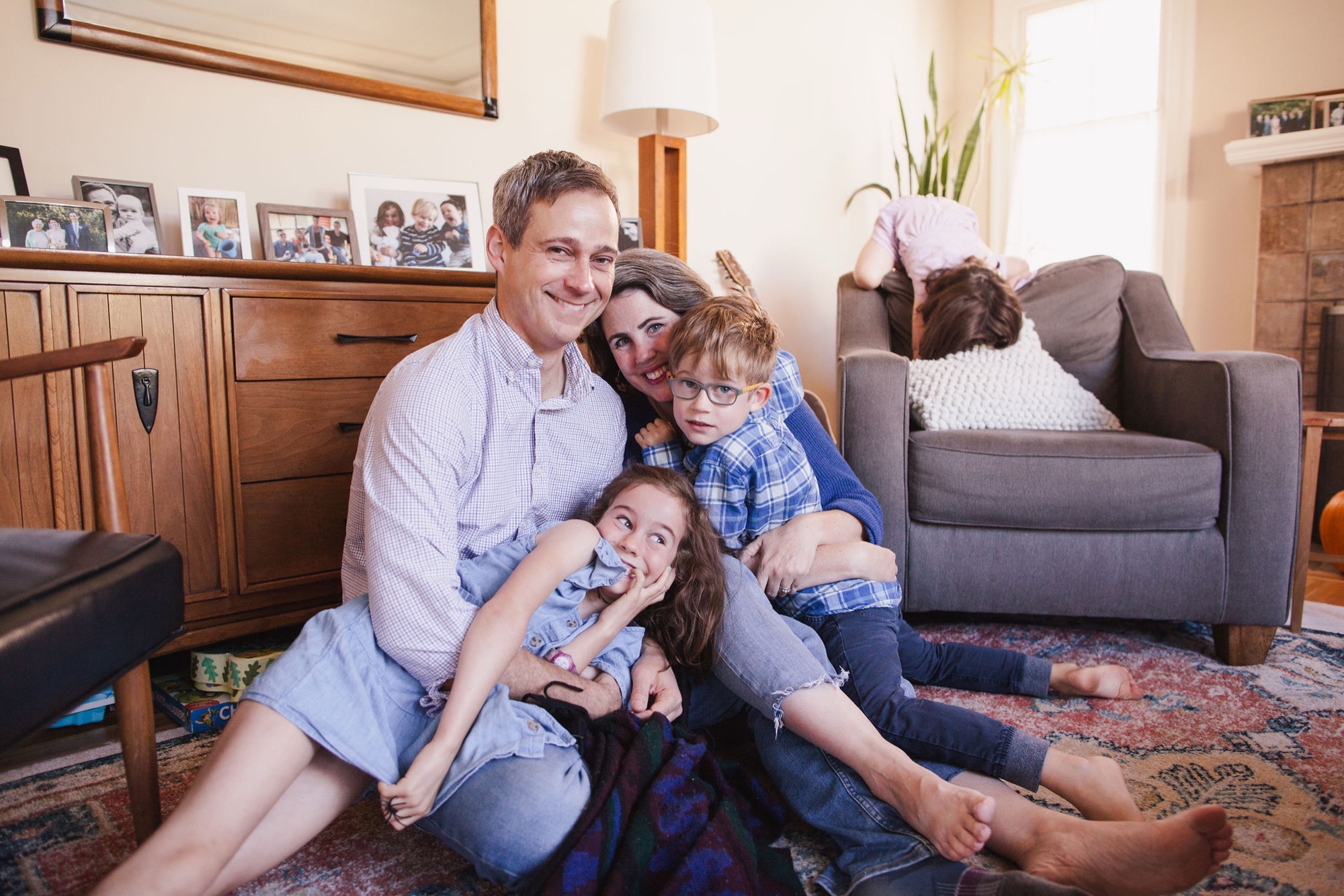 A family of four laughs together on a living room floor, bathed in warm natural light streaming through windows, creating an intimate moment of genuine joy and connection