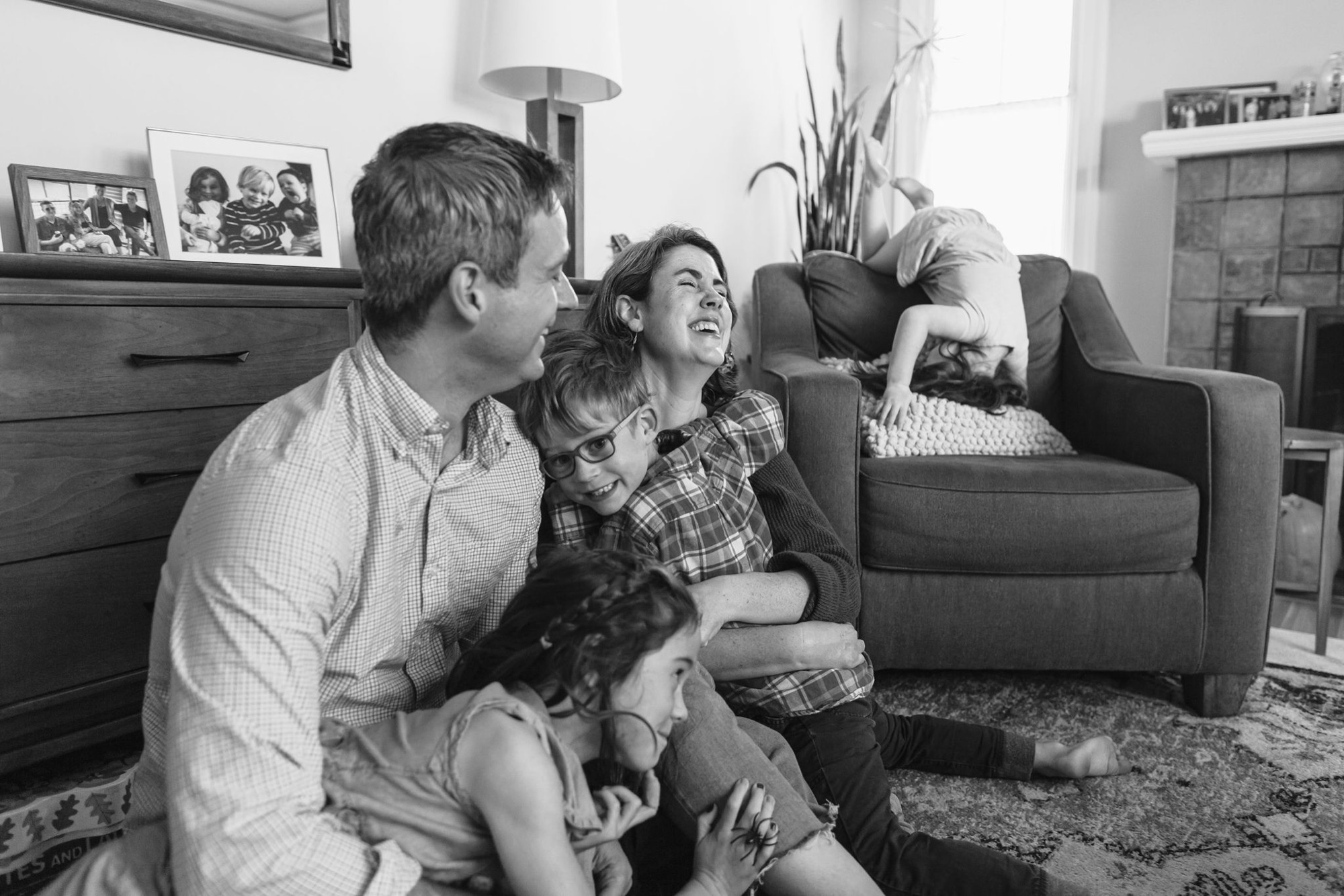Family of five sits together on living room floor, laughing upward as youngest child playfully drapes over the couch arm behind them