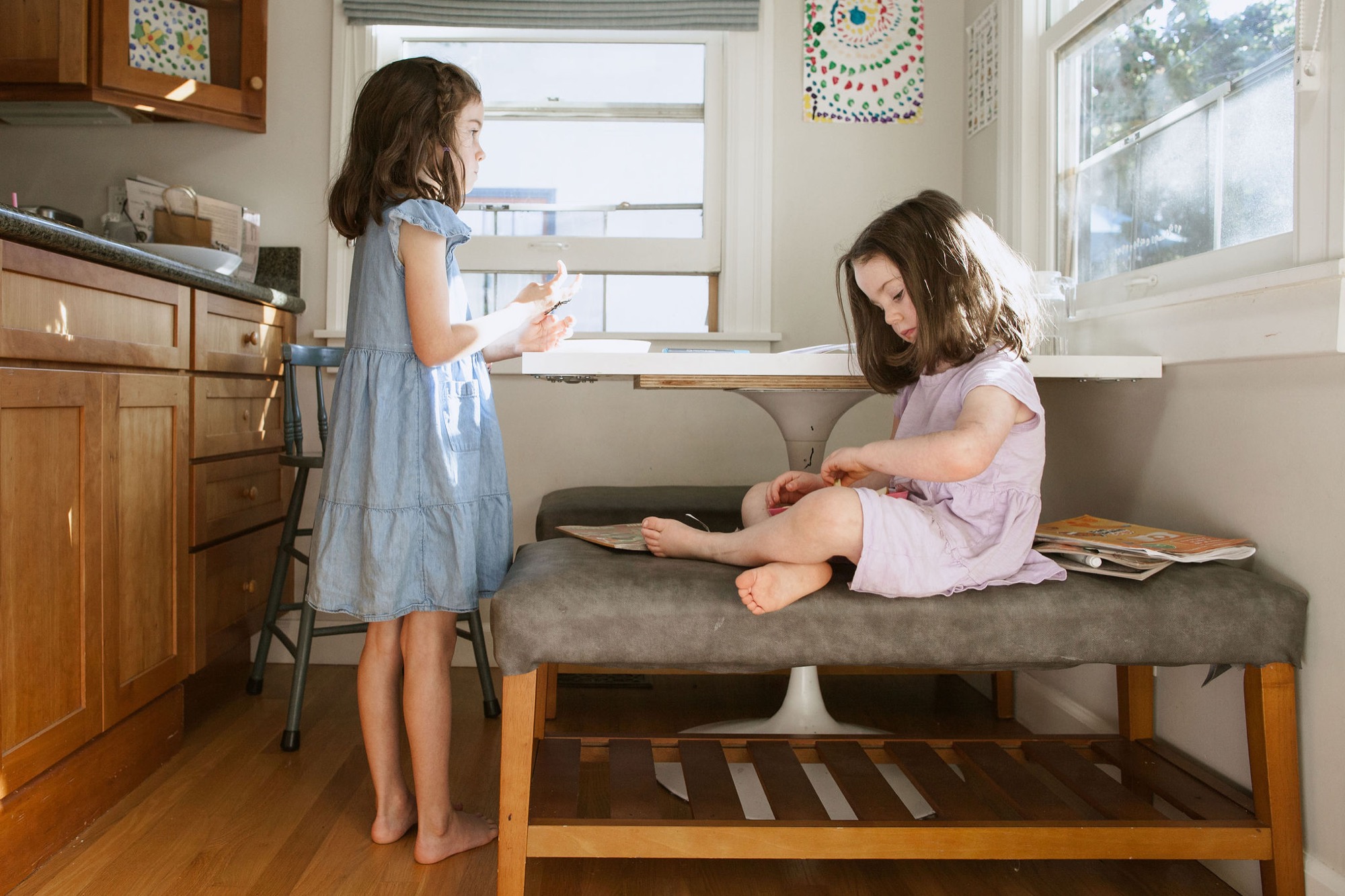 Two girls sit on a mid-century bench in bright kitchen light, the standing child gesturing animatedly while her sister reads below