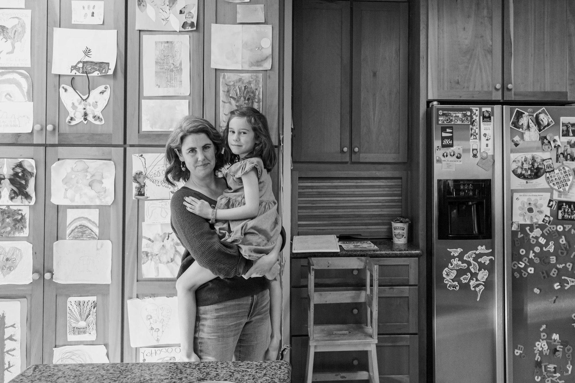 A mother holds her young daughter in a kitchen studio, both gazing thoughtfully toward the camera while surrounded by children's artwork pinned to glass cabinet doors