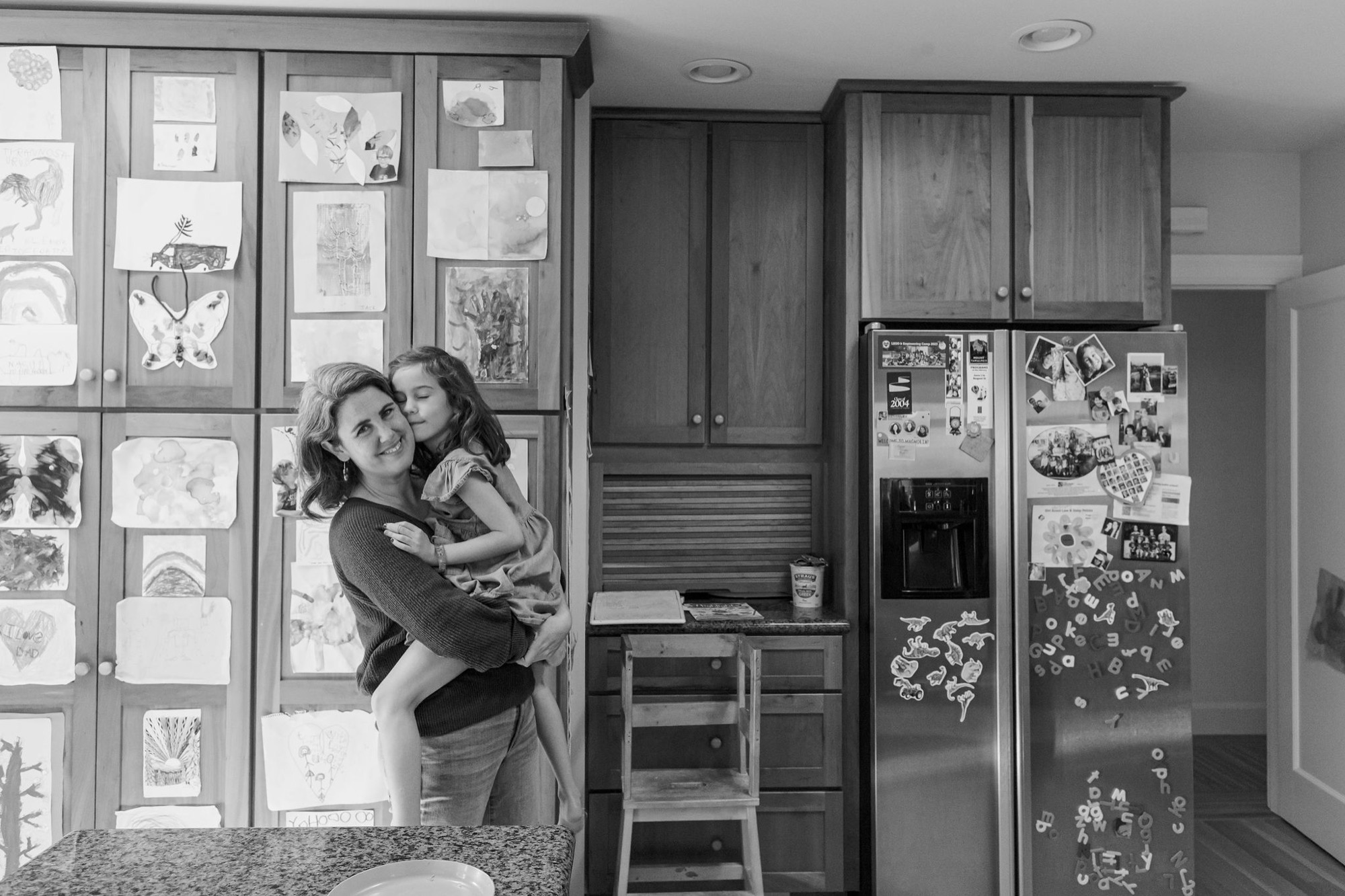 A mother holds her young daughter in a modern kitchen, both gazing warmly at each other beside a refrigerator covered with children's artwork and family memories