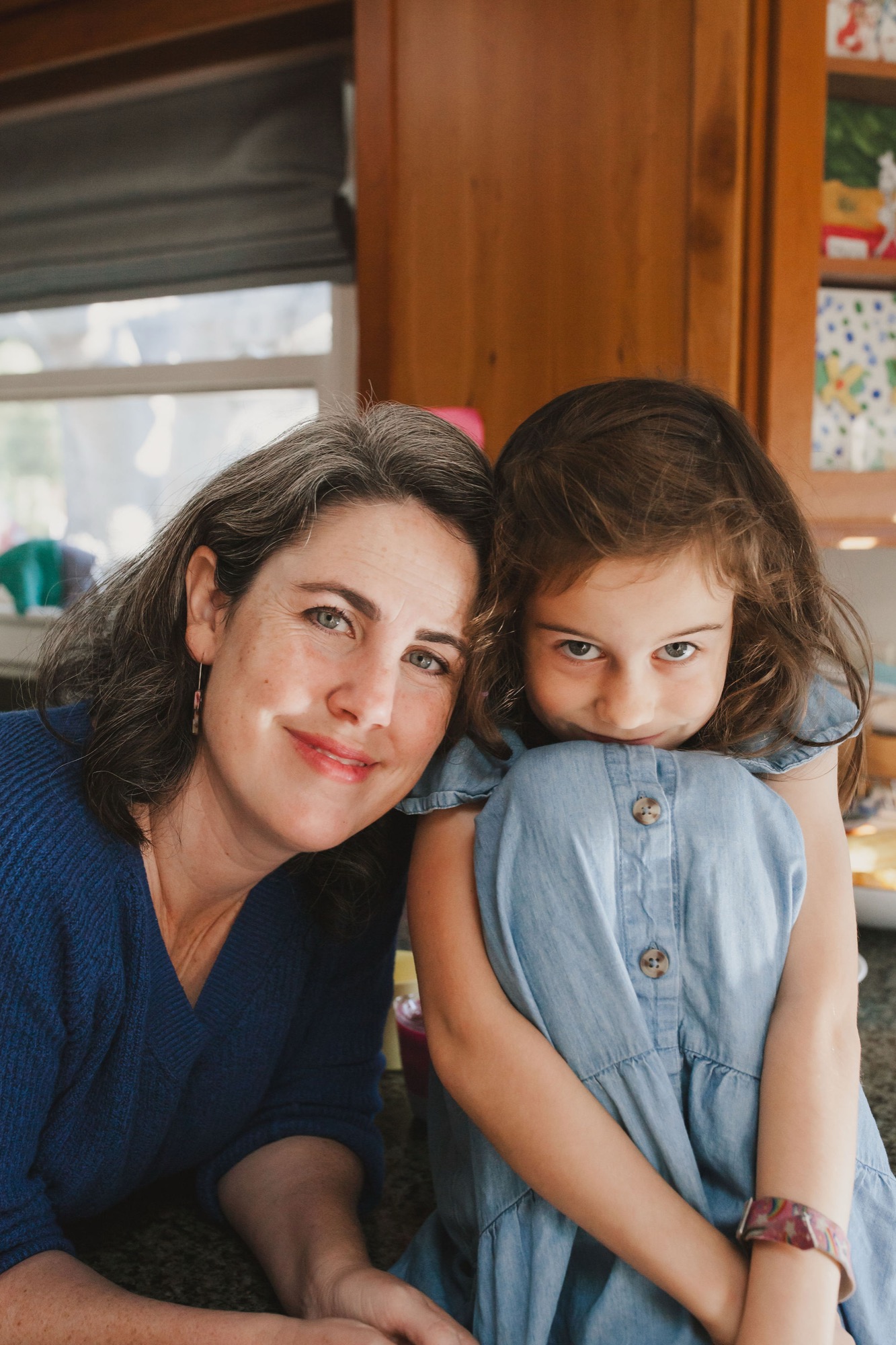 A mother and daughter sit close together indoors, their warm expressions and relaxed posture conveying genuine affection in soft, natural window light