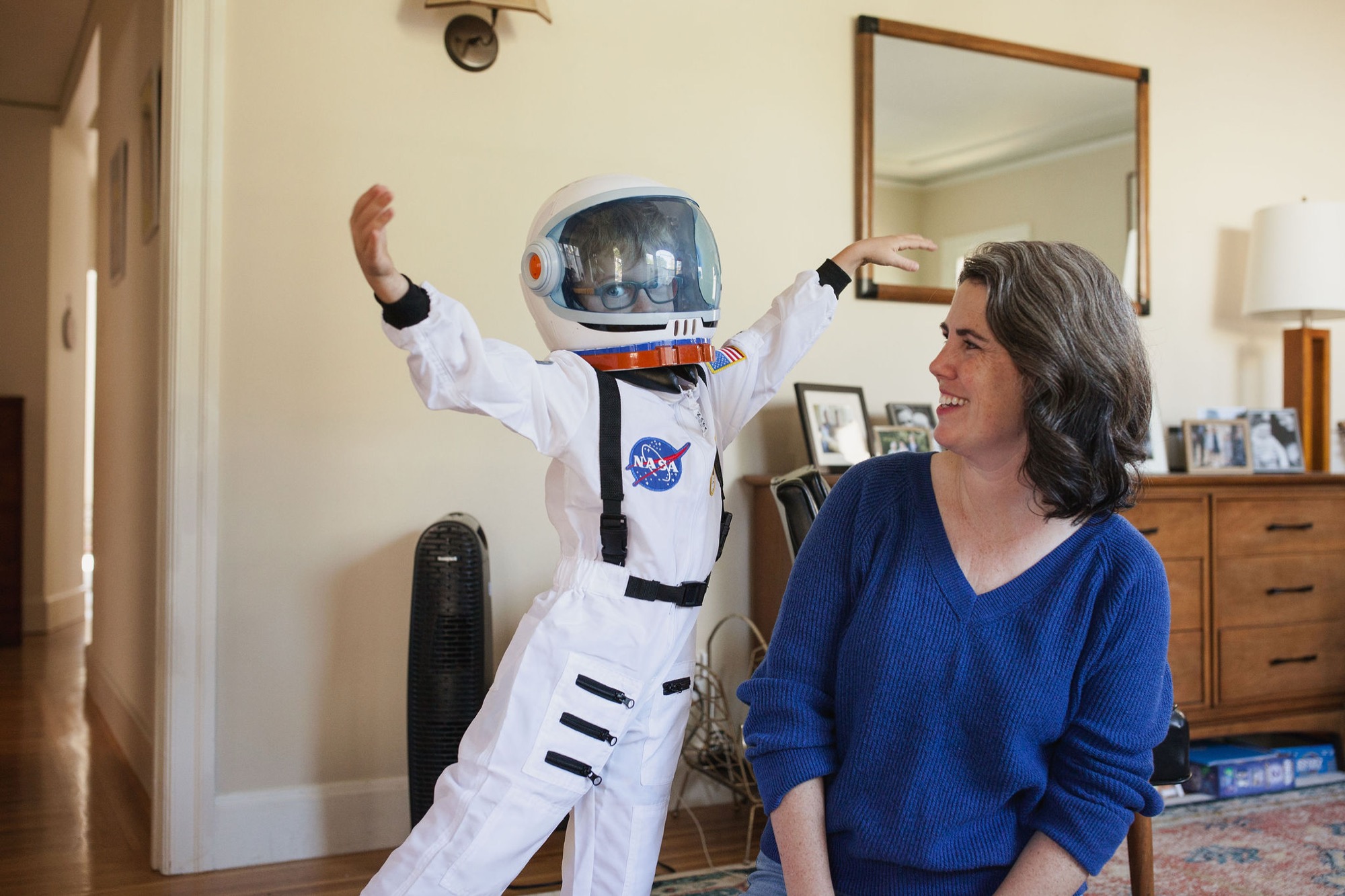 A woman in a blue sweater smiles warmly at a NASA astronaut robot figure with arms raised triumphantly in a home interior