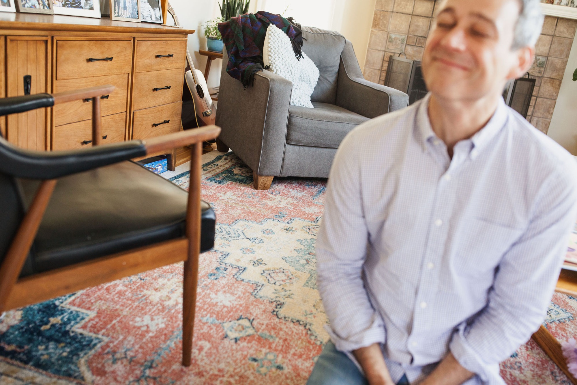 Man in lavender shirt sits thoughtfully in a sunlit living room among mid-century modern furniture and a patterned rug