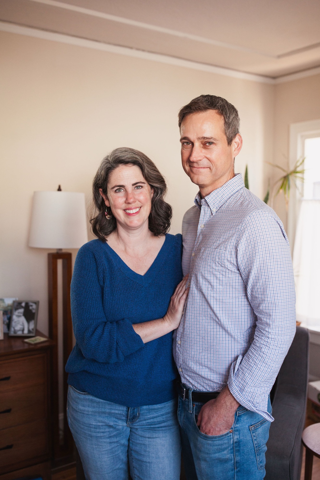 A couple smiles warmly at the camera in their living room, with soft natural light illuminating their relaxed, affectionate poses near wooden furniture
