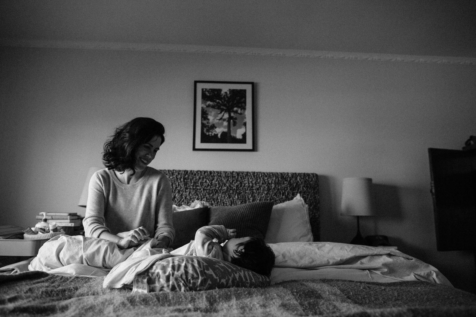 A woman sits cross-legged on a bed, smiling down at a child lying beside her, soft natural light illuminating their intimate moment