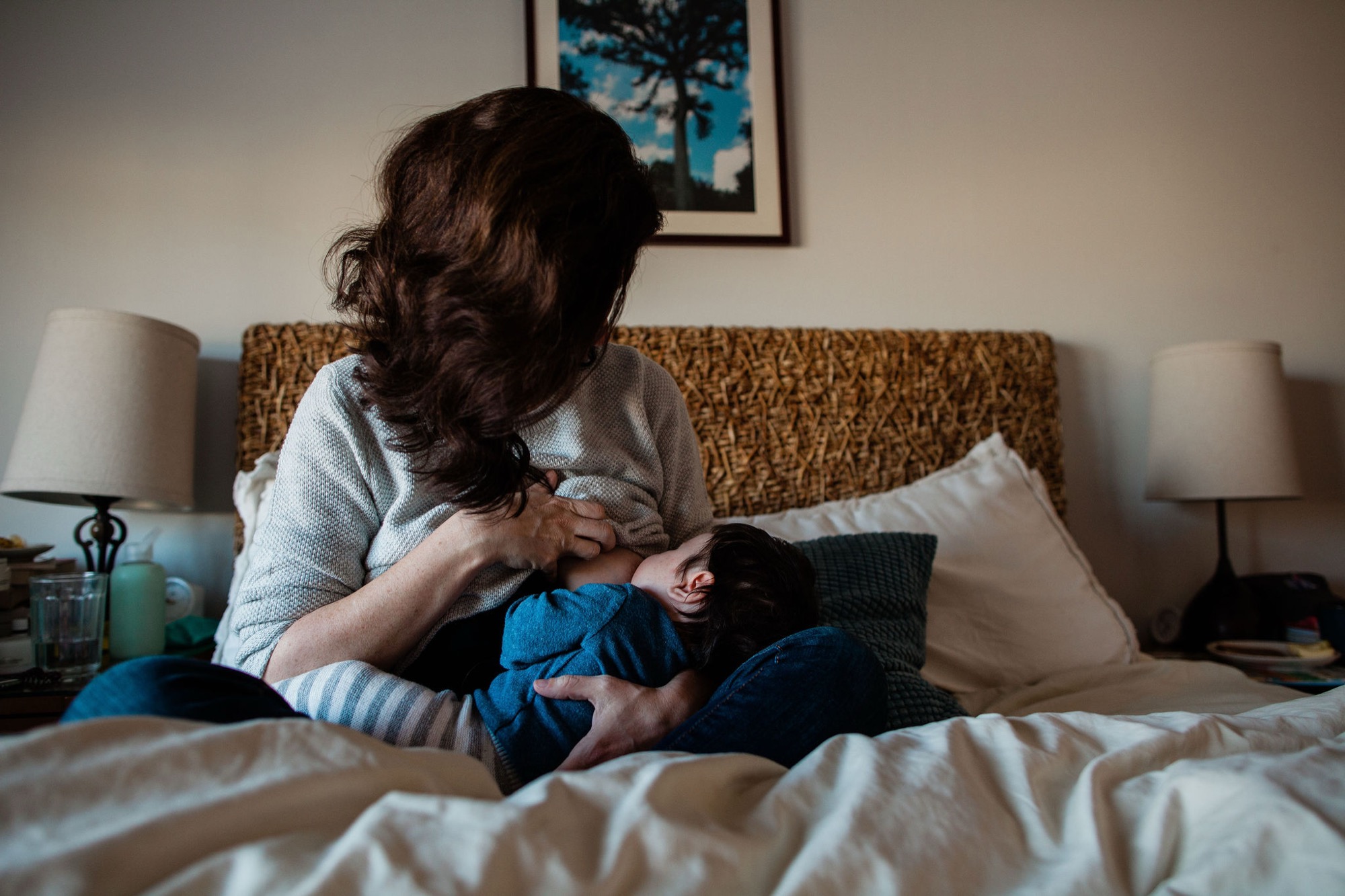 Mother gazes down tenderly at her infant son cradled in her arms on a textured bed, soft bedside lamps flanking the intimate moment