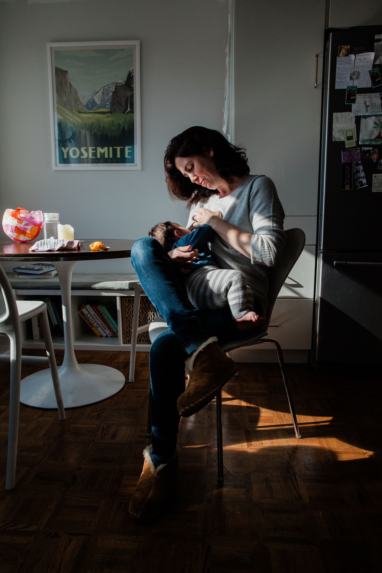 A woman in a striped cardigan holds her toddler on her lap while seated, both bathed in warm afternoon sunlight streaming across the wooden floor
