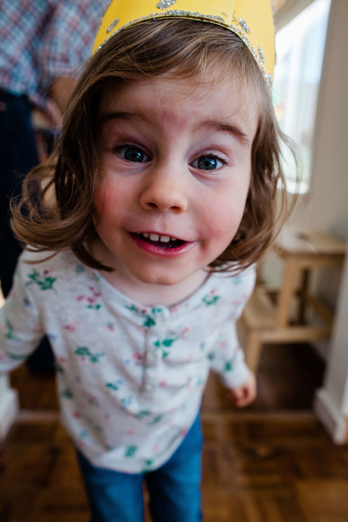 A delighted toddler in a yellow crown and floral shirt gazes up with wide eyes and an open-mouthed smile, captured from above in warm indoor light