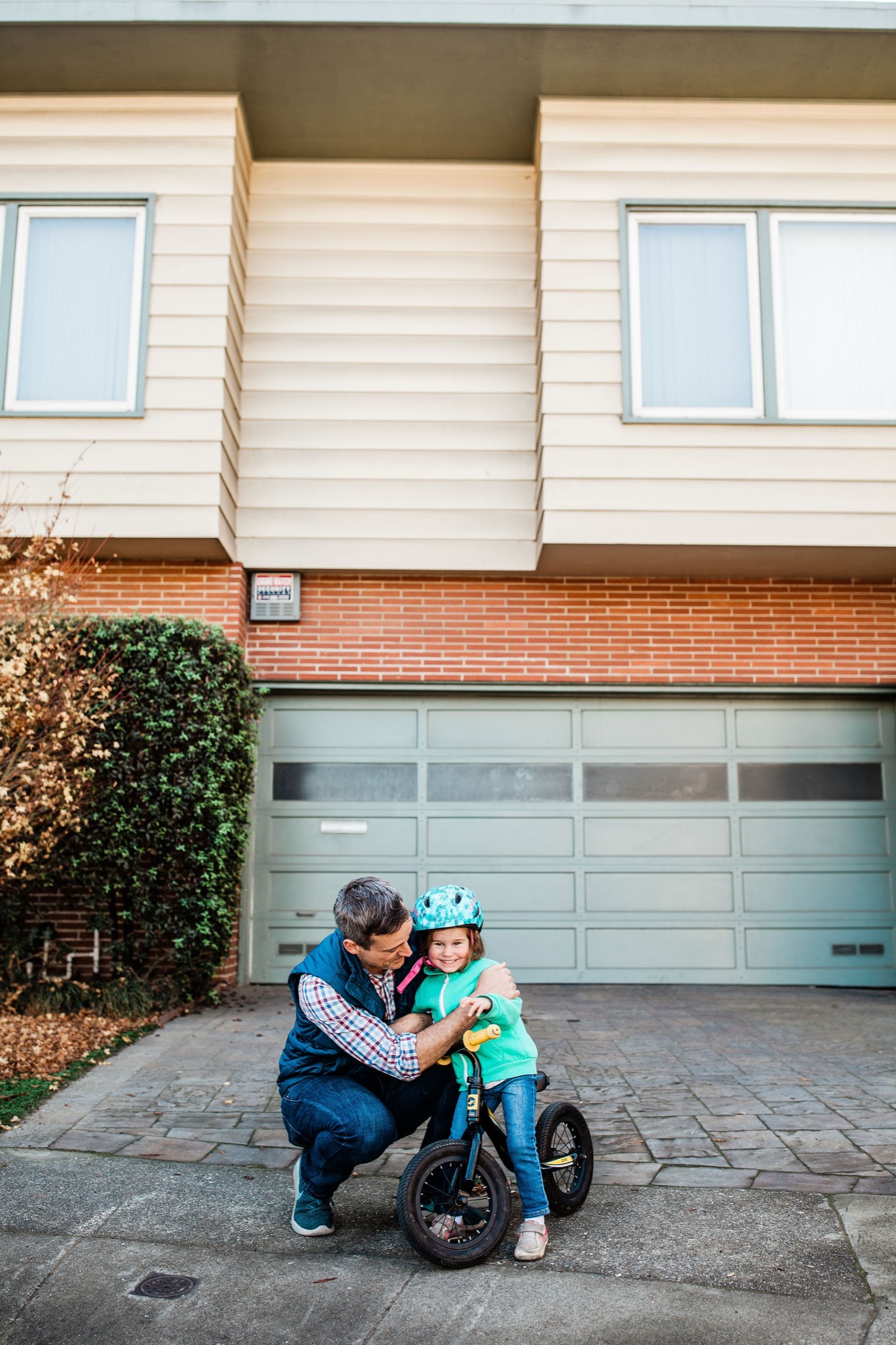 A father crouches beside his young daughter on her balance bike, both beaming with joy on their driveway in afternoon light