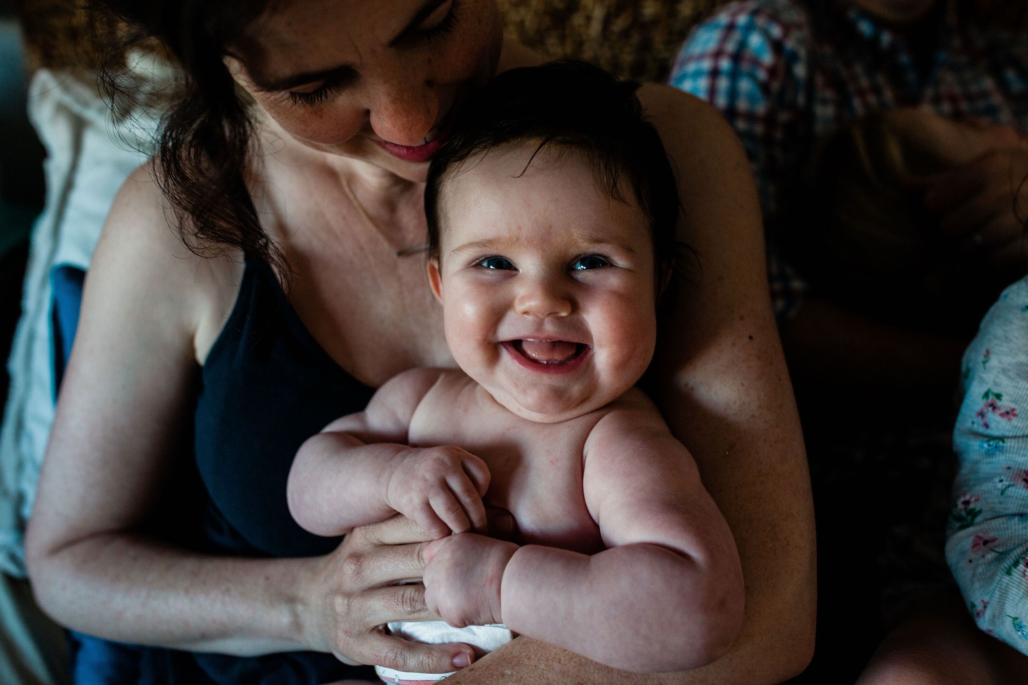 A mother holds her laughing baby close, both beaming with genuine joy as warm natural light softly illuminates their faces