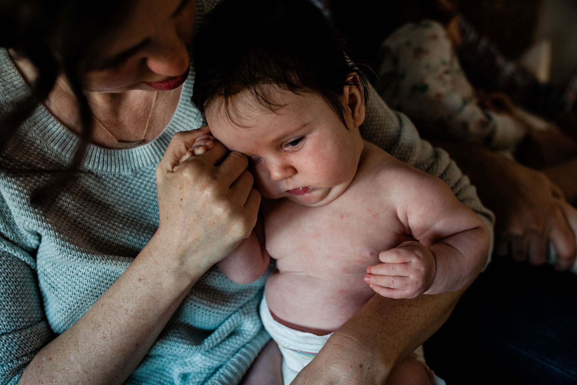 A parent tenderly holds their newborn in soft indoor light, the baby's hand reaching toward their chest while both gaze downward in quiet intimacy