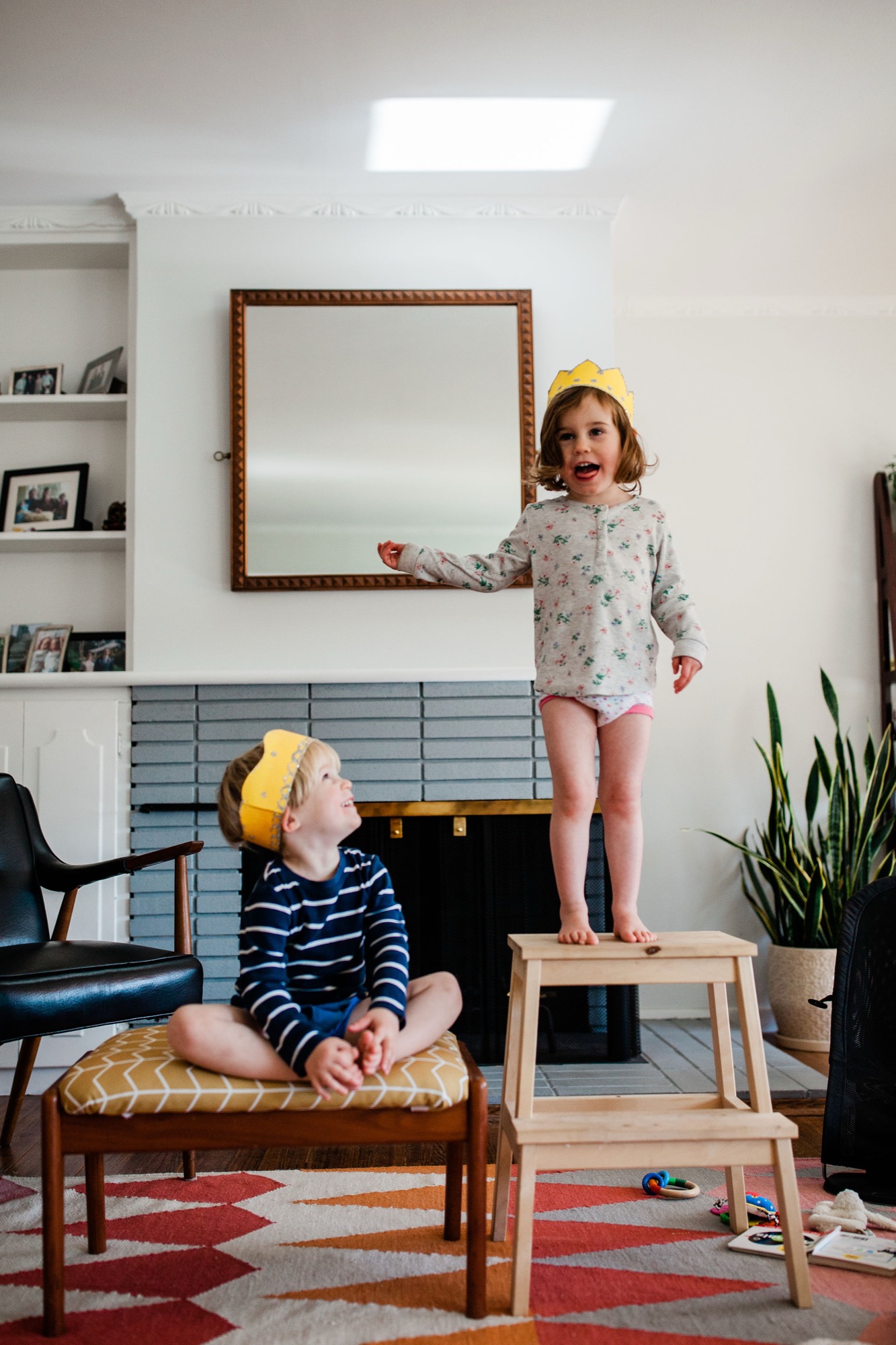 Two young children wearing paper crowns play together in a bright living room, the girl standing on a wooden step stool while her brother sits on a cushioned bench below, both smiling with pure joy