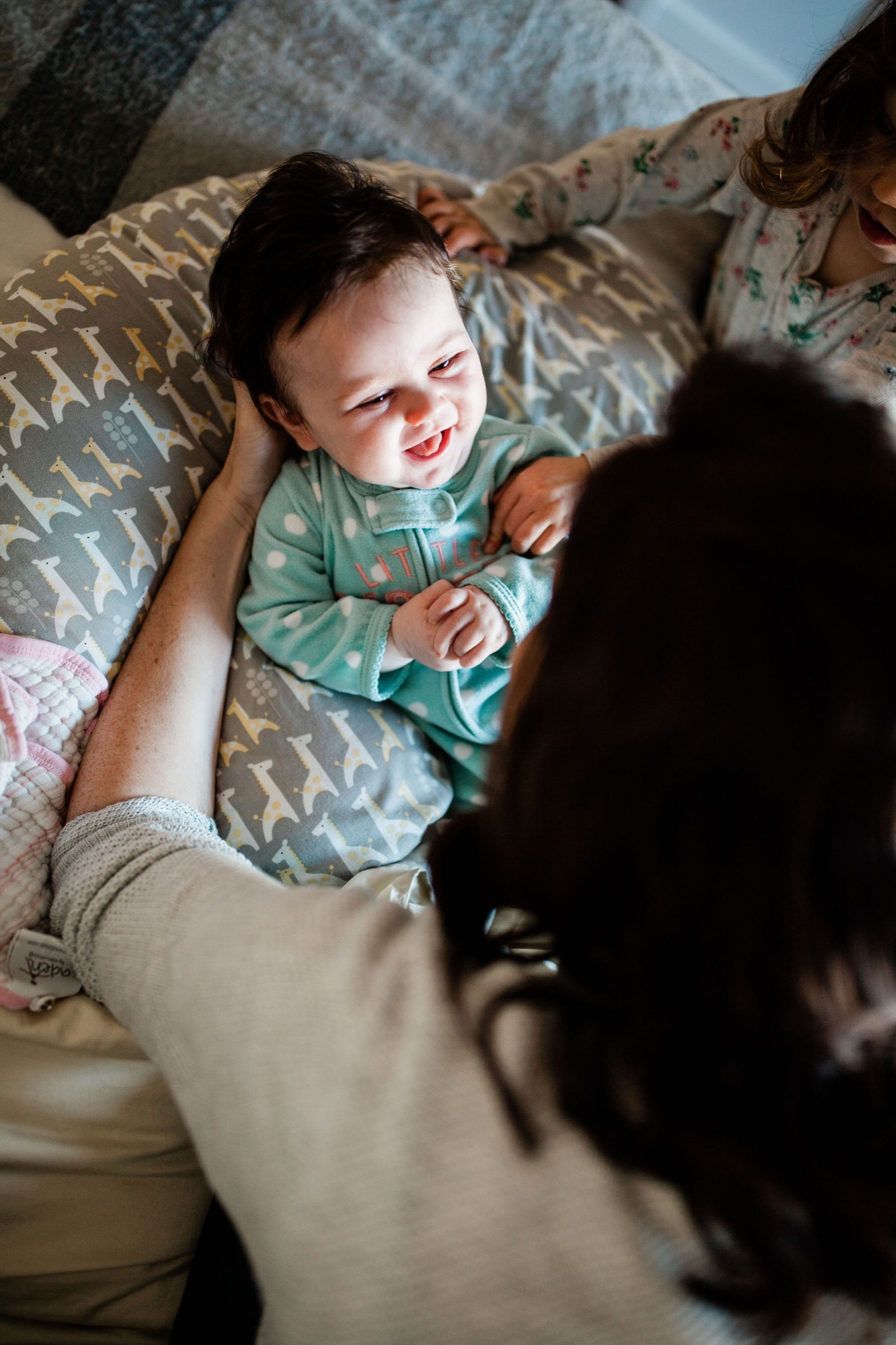 A delighted baby in a green shirt beams while being held by a parent on a patterned bed, with natural light streaming across the intimate moment
