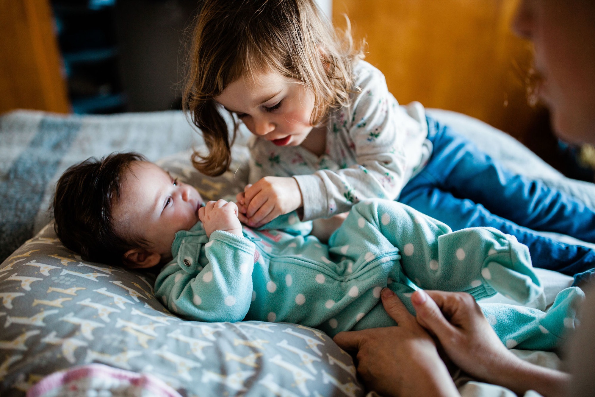Older sister gazes down tenderly at her newborn sibling lying on patterned bedding, their hands gently touching in warm natural light