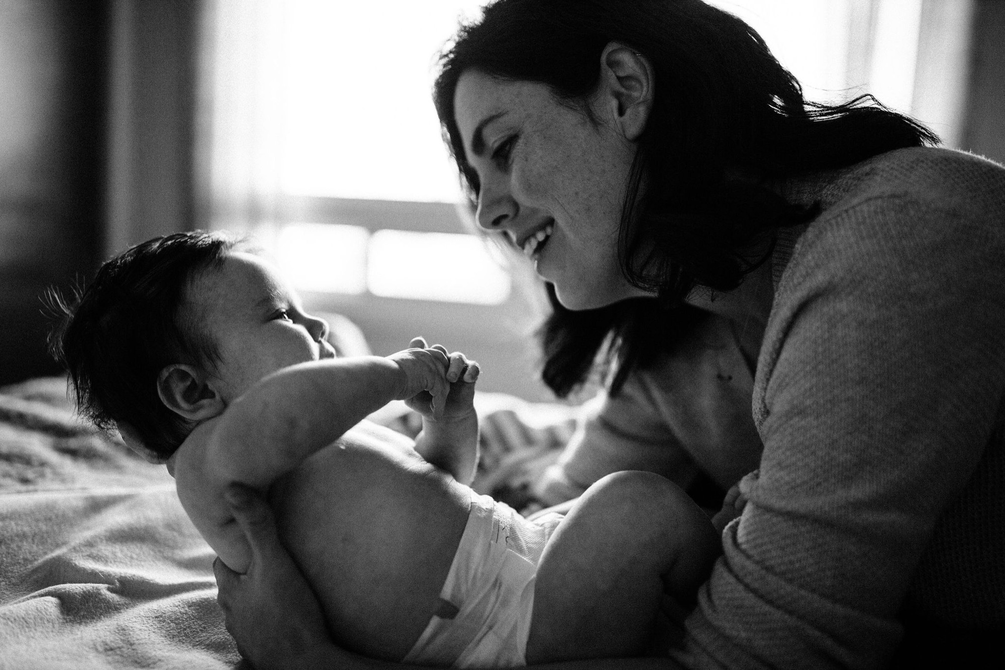 A mother gazes down at her infant son with a genuine smile as he reaches toward her face, bathed in soft window light