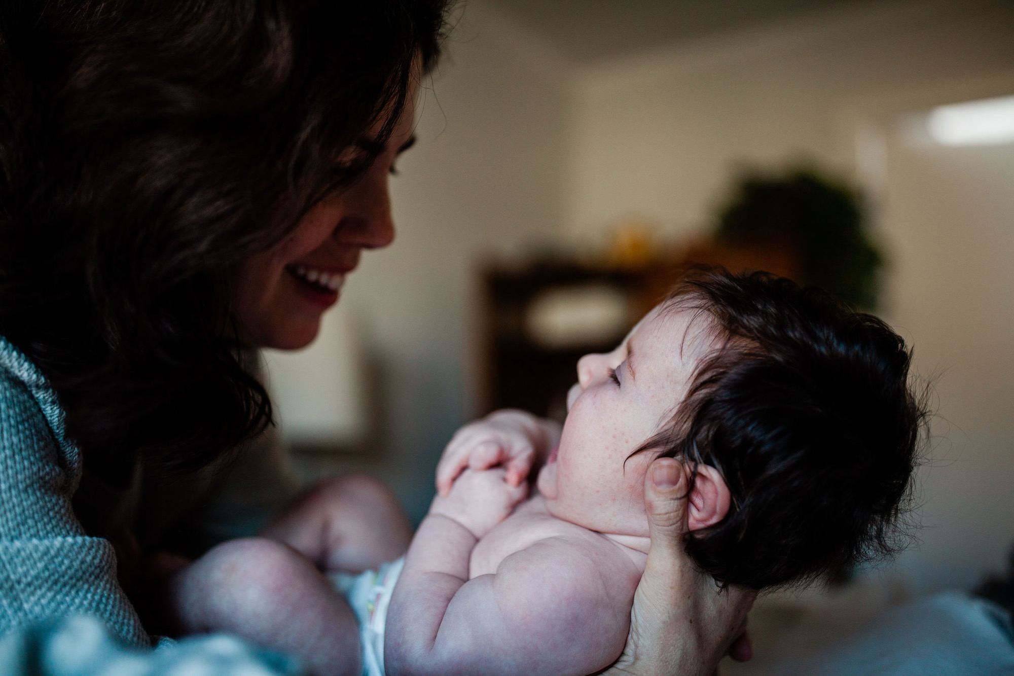 A mother in a teal sweater gazes down at her newborn with a genuine smile, cradling the sleeping infant in soft, warm indoor light