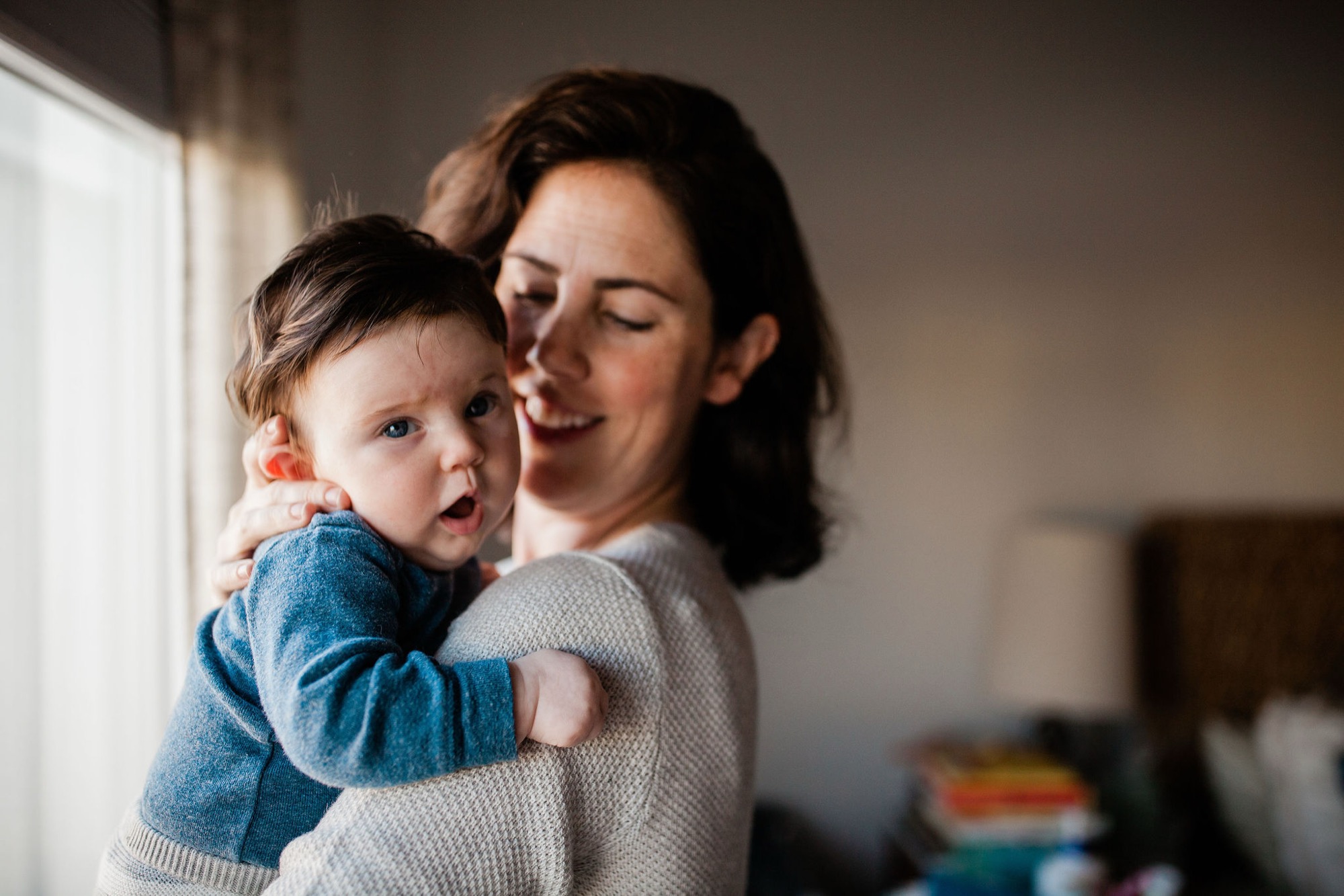 A woman in a cream sweater holds her toddler in a blue jacket near a bright window, both gazing with genuine joy and wonder