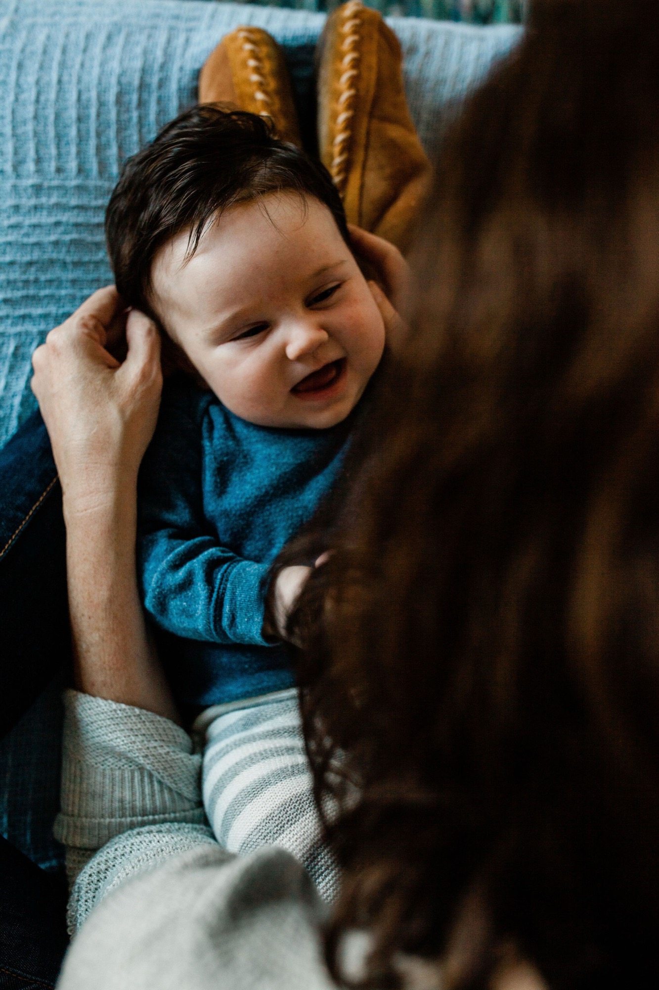 A joyful infant in a blue shirt laughs while being held by an adult on a light blue textured couch, with warm natural light illuminating the tender moment